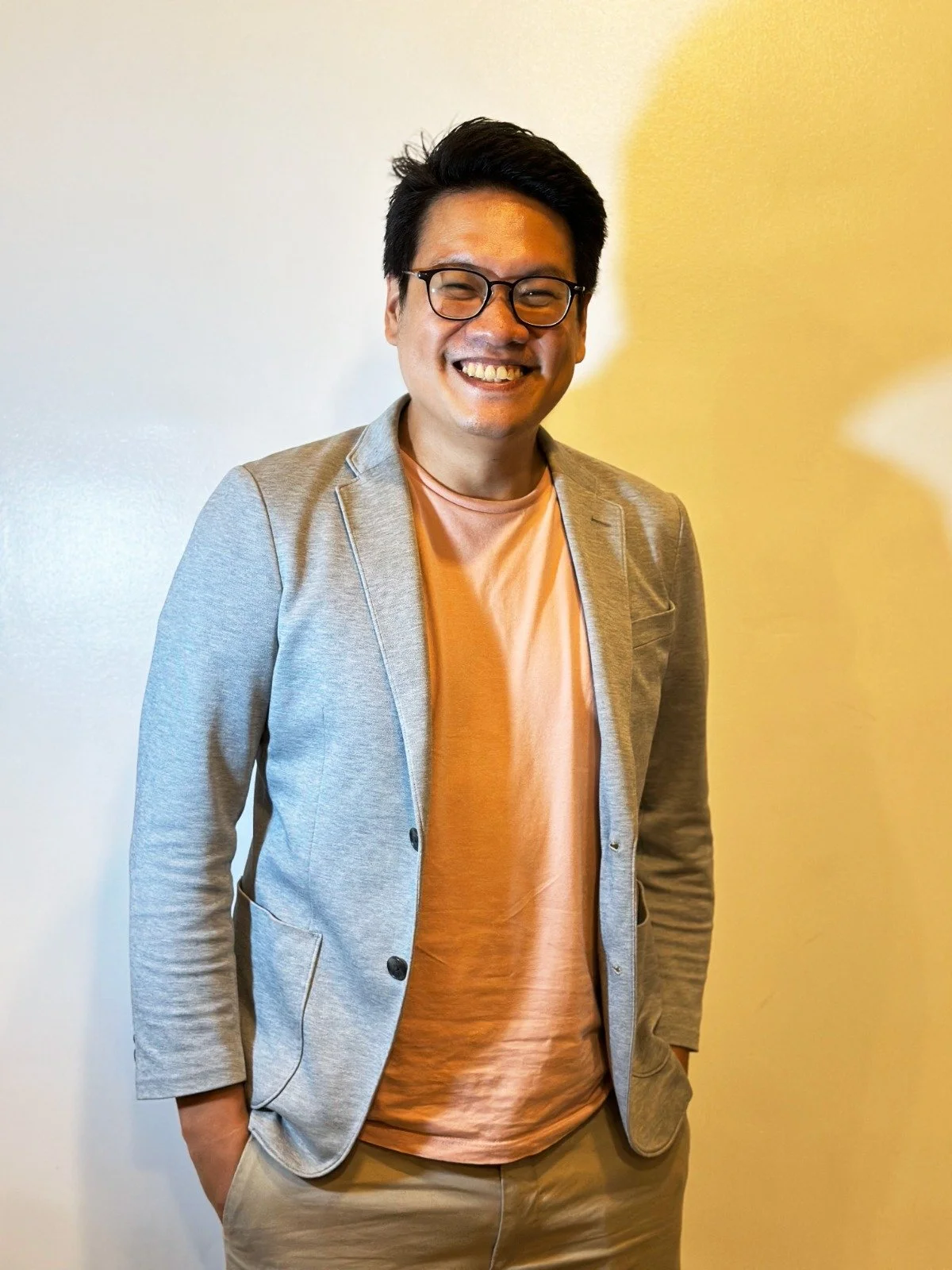 Faction author Lio Mangubat. Here he is pictured with wearing black glasses, a light gray blazer, and beige pants standing against a plain wall.