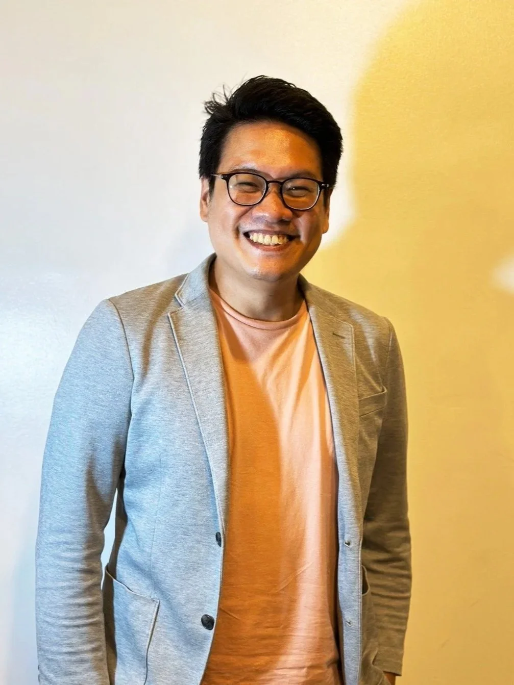 Faction author Lio Mangubat. Here he is pictured with wearing black glasses, a light gray blazer, and beige pants standing against a plain wall.