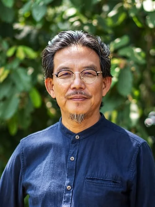Faction author Ooi Kee Beng. Here he is pictured with glasses and a goatee, wearing a dark blue button-up shirt, standing outdoors with green foliage in the background.