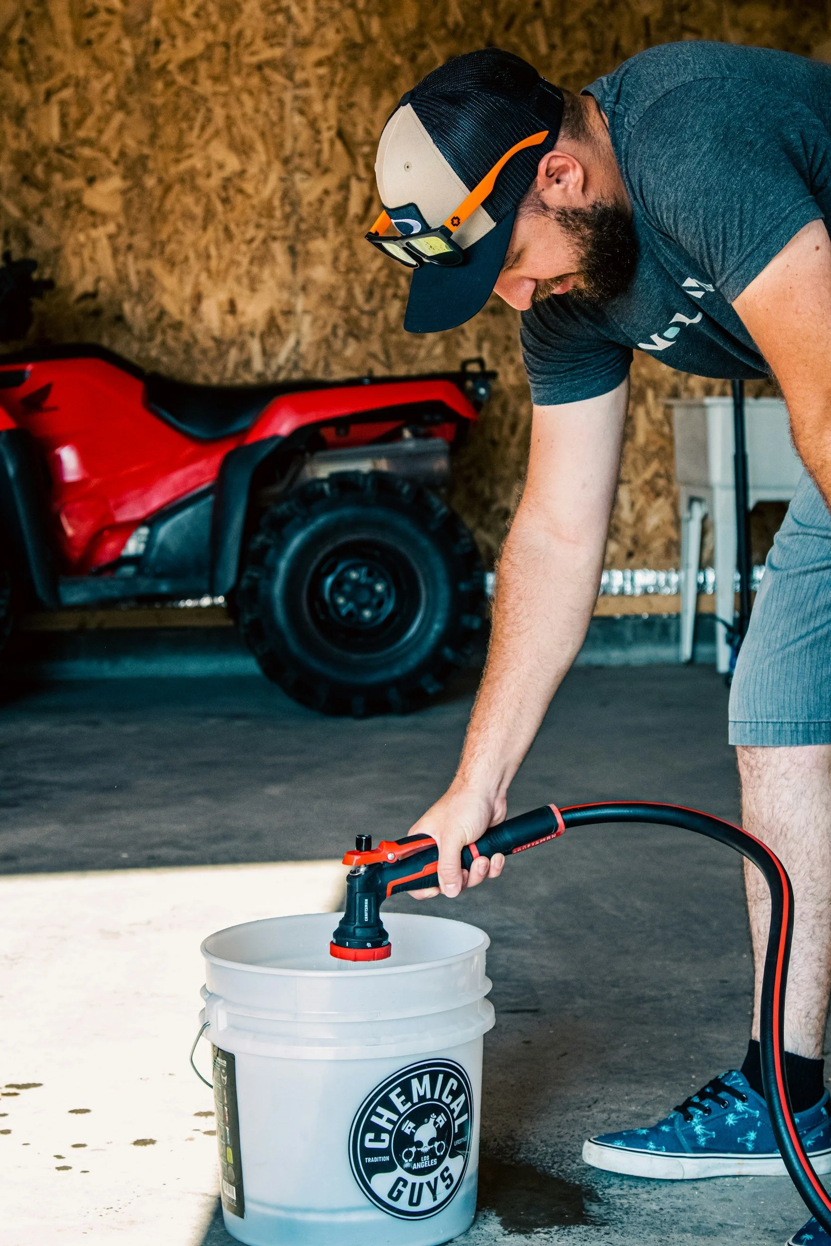 A man wearing a black cap, glasses, and a dark gray T-shirt with a logo is using a cordless heat gun to heat a white plastic bucket in a garage with a wooden wall, a red all-terrain vehicle, and storage shelves in the background.