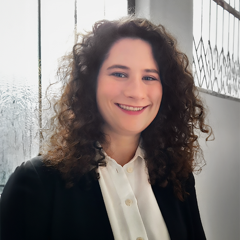 A young woman with curly brown hair smiling, wearing a white blouse and black blazer, standing next to a window with raindrops and a wire fence outside.