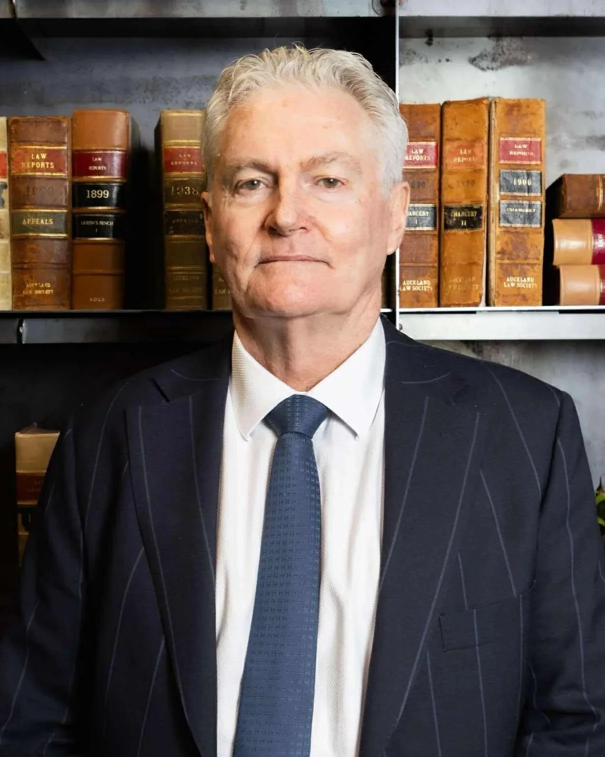 A middle-aged man with gray hair in a dark pinstripe suit and blue tie, standing in front of a shelf filled with old law books.