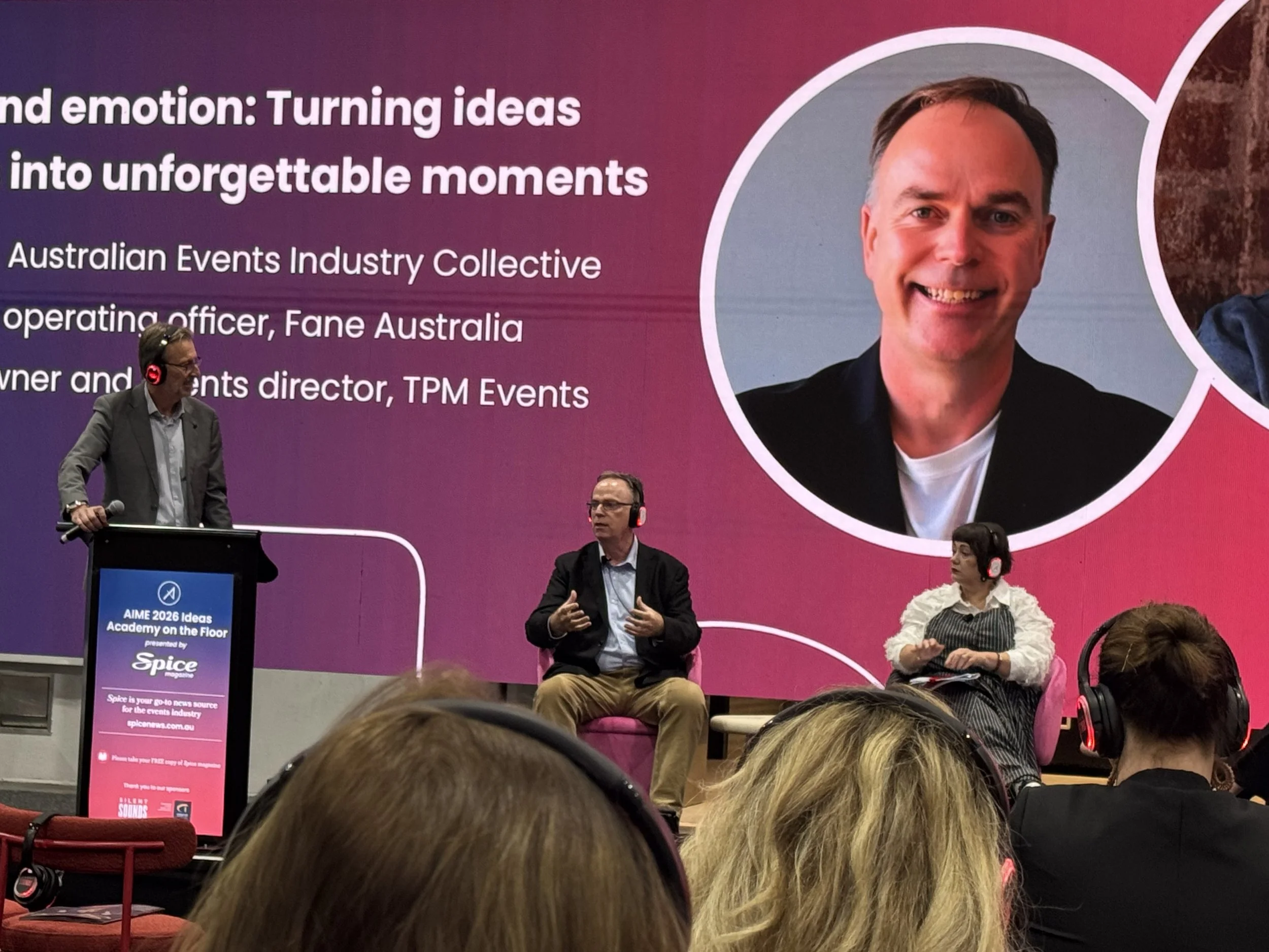 A panel discussion at a conference with several people seated on stage, a large pink and purple backdrop displaying text about turning ideas into unforgettable moments, and a few audience members visible in the foreground.