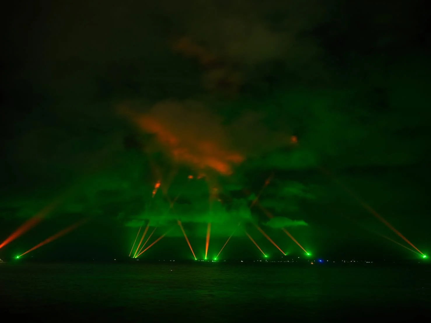 Nighttime scene of a laser light show over a body of water, with green and red laser beams shooting into the sky and illuminating clouds.