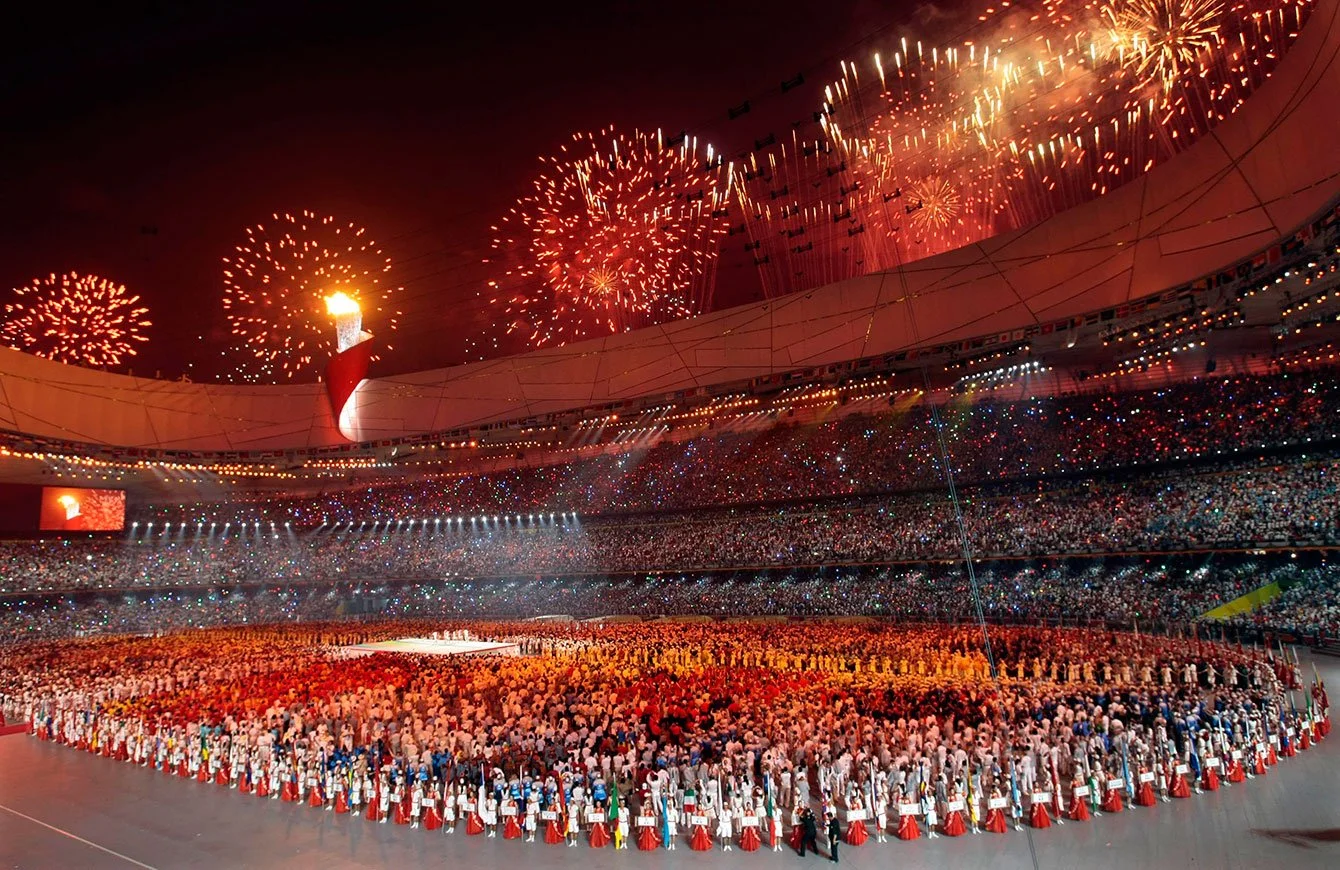 Large crowd in a stadium celebrating with fireworks and torch lighting during an Olympic event.