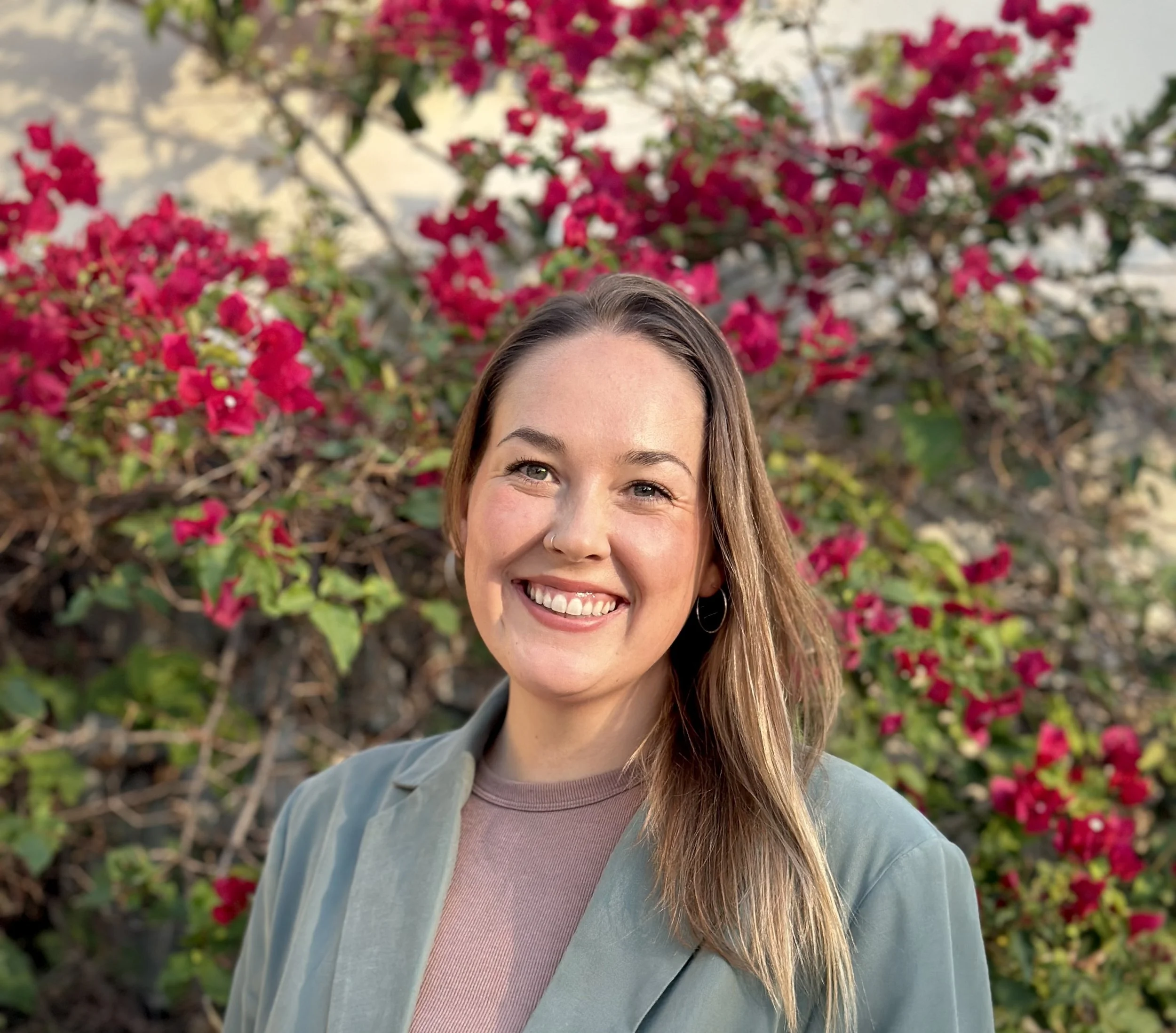 A young woman smiling outdoors in front of pink flowering bushes.