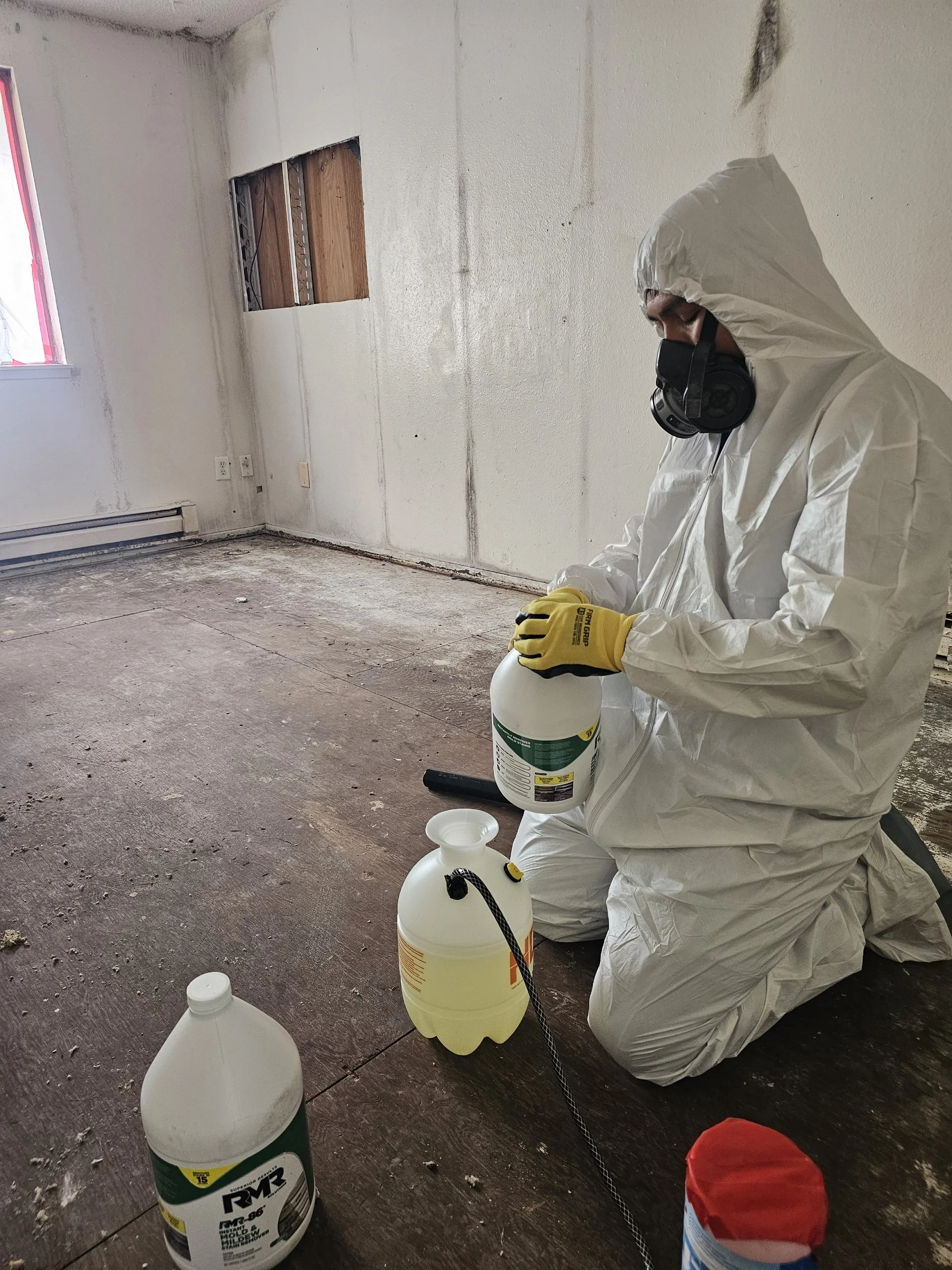 A person dressed in a hazmat suit, kneeling, wearing a mask and gloves, holding a container of liquid. They are in a partially renovated or damaged room with exposed wall wiring, missing wall panel, and construction materials, including a large bottle and a small container on the floor.