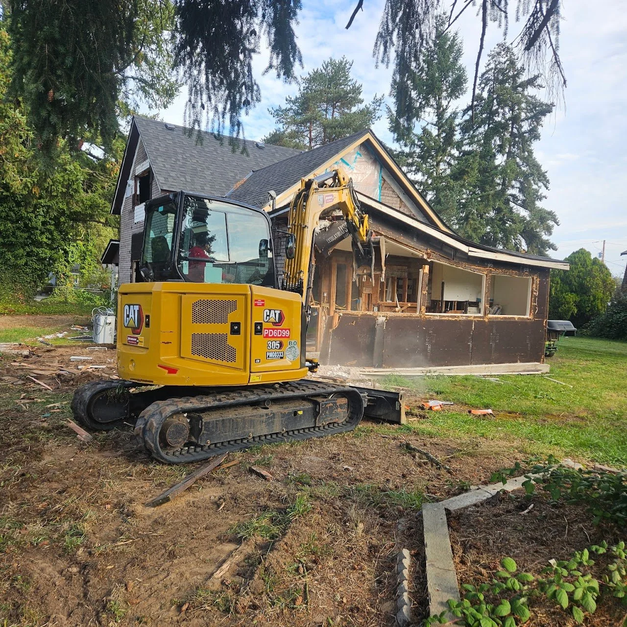 A yellow Caterpillar mini excavator demolishing a small house, with part of the structure and walls visibly torn down, on a grassy yard with trees in the background.