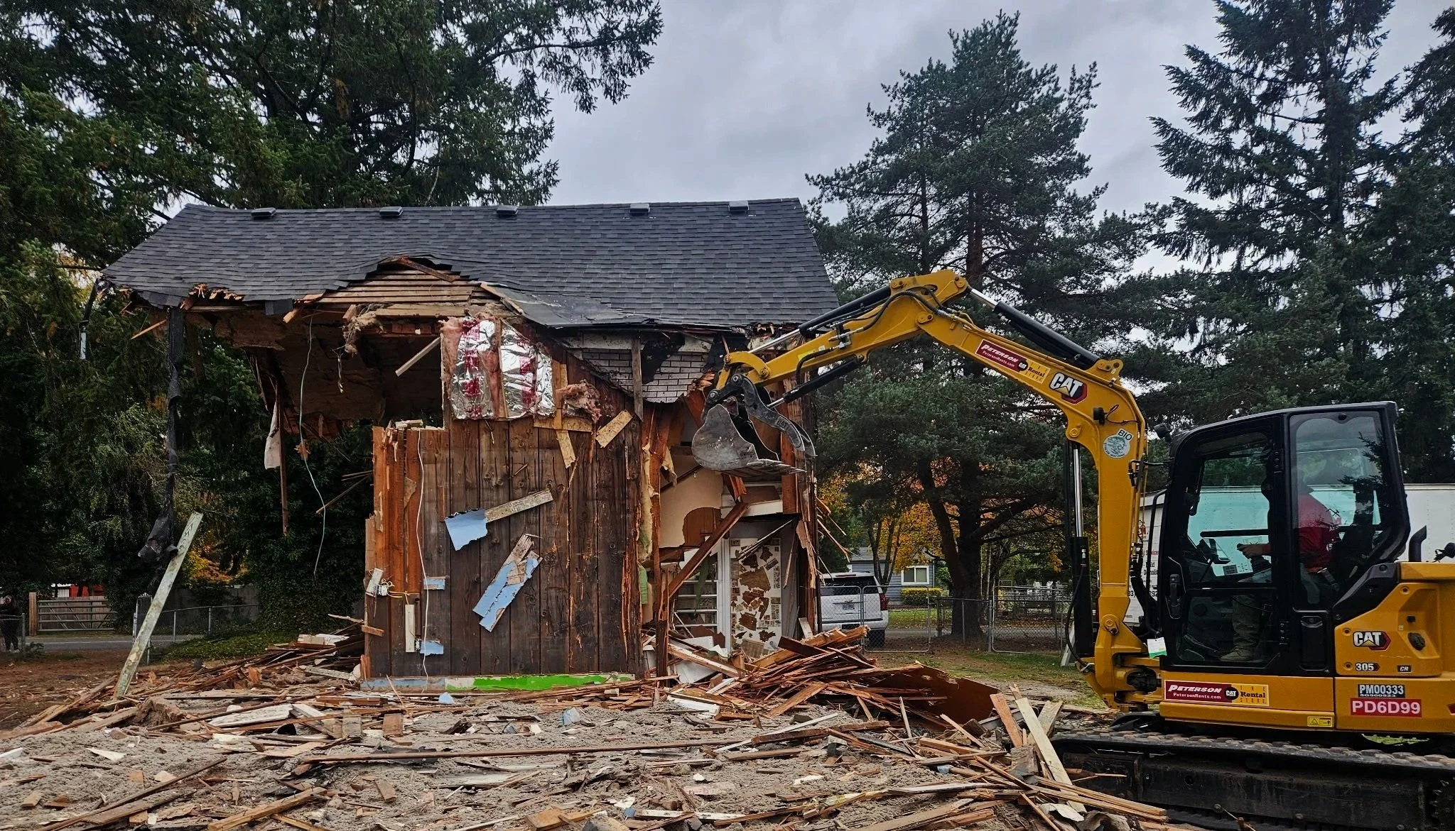 A partially demolished wooden house with damaged roof and walls, surrounded by debris, with a yellow excavator nearby and trees in the background.