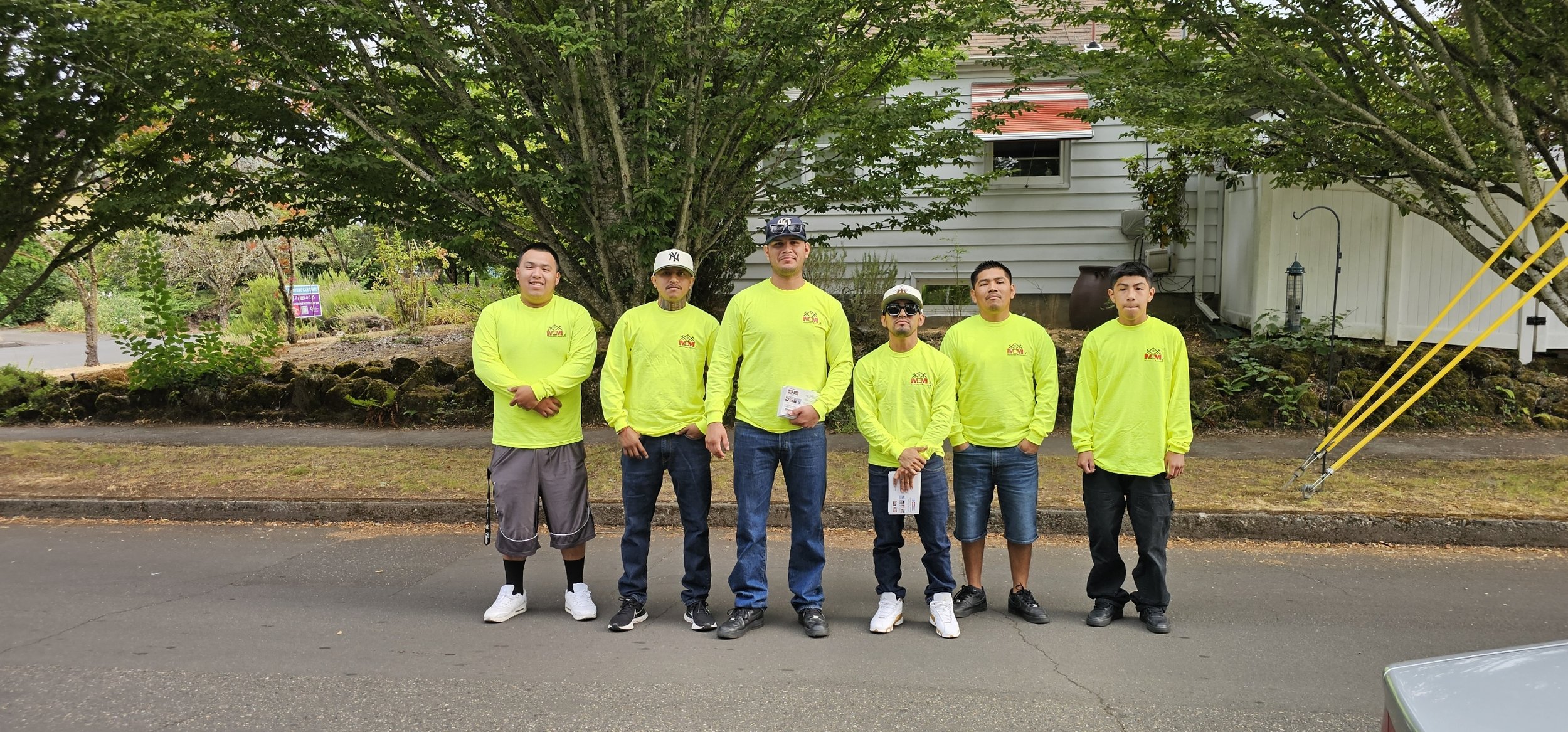 Six men standing side by side on a street, wearing matching bright yellow long-sleeve shirts with a logo, with trees and a house in the background.