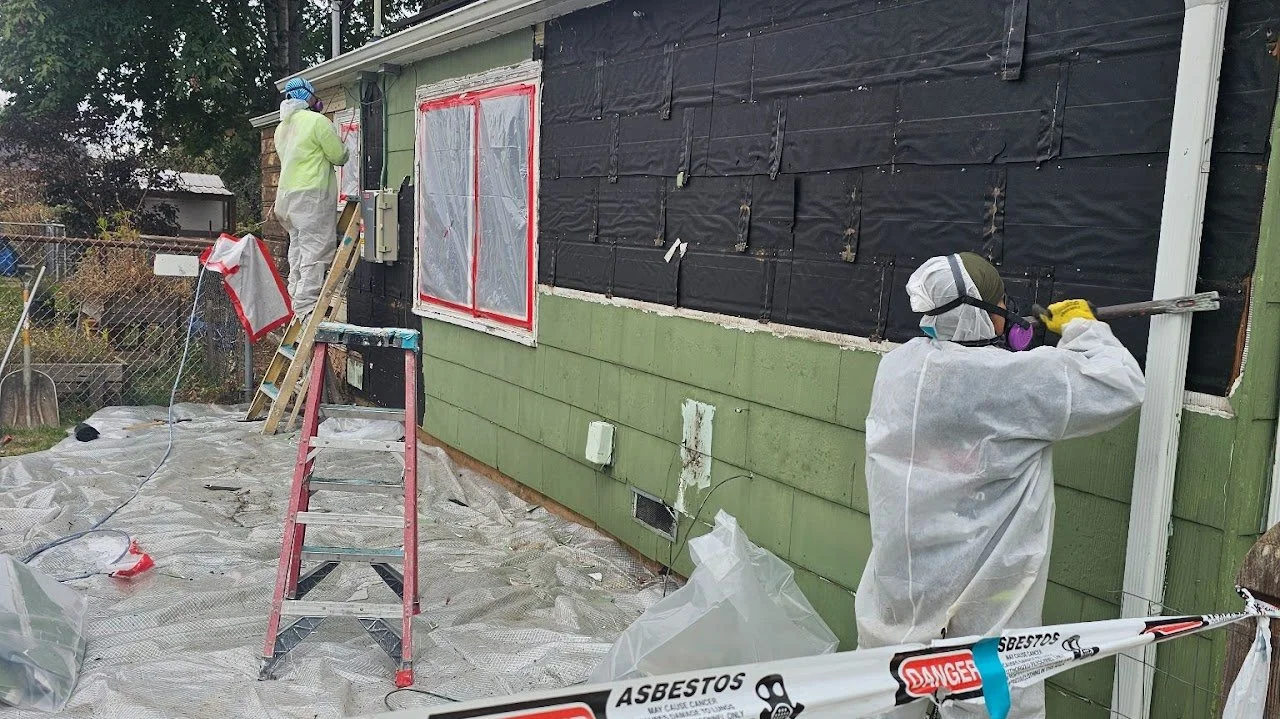 Two workers in protective suits and masks removing asbestos from the exterior wall of a house, which is partially covered with black asbestos siding and plastic sheeting, on a backyard deck with a ladder, caution tape, and tools present.