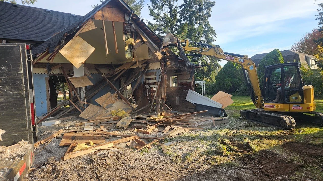 A small excavator demolishing the side of a house, with debris and broken wood scattered on the ground.