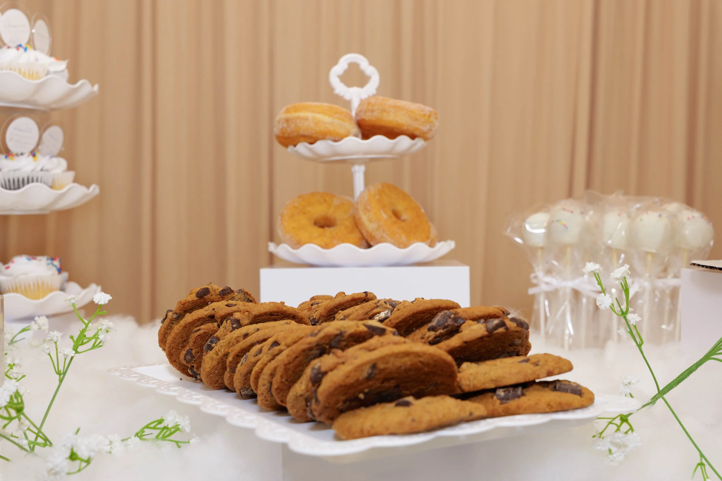 Display of chocolate chip cookies on a white tray, surrounded by small white flowers, with a tiered stand of donuts and cupcakes in the background.
