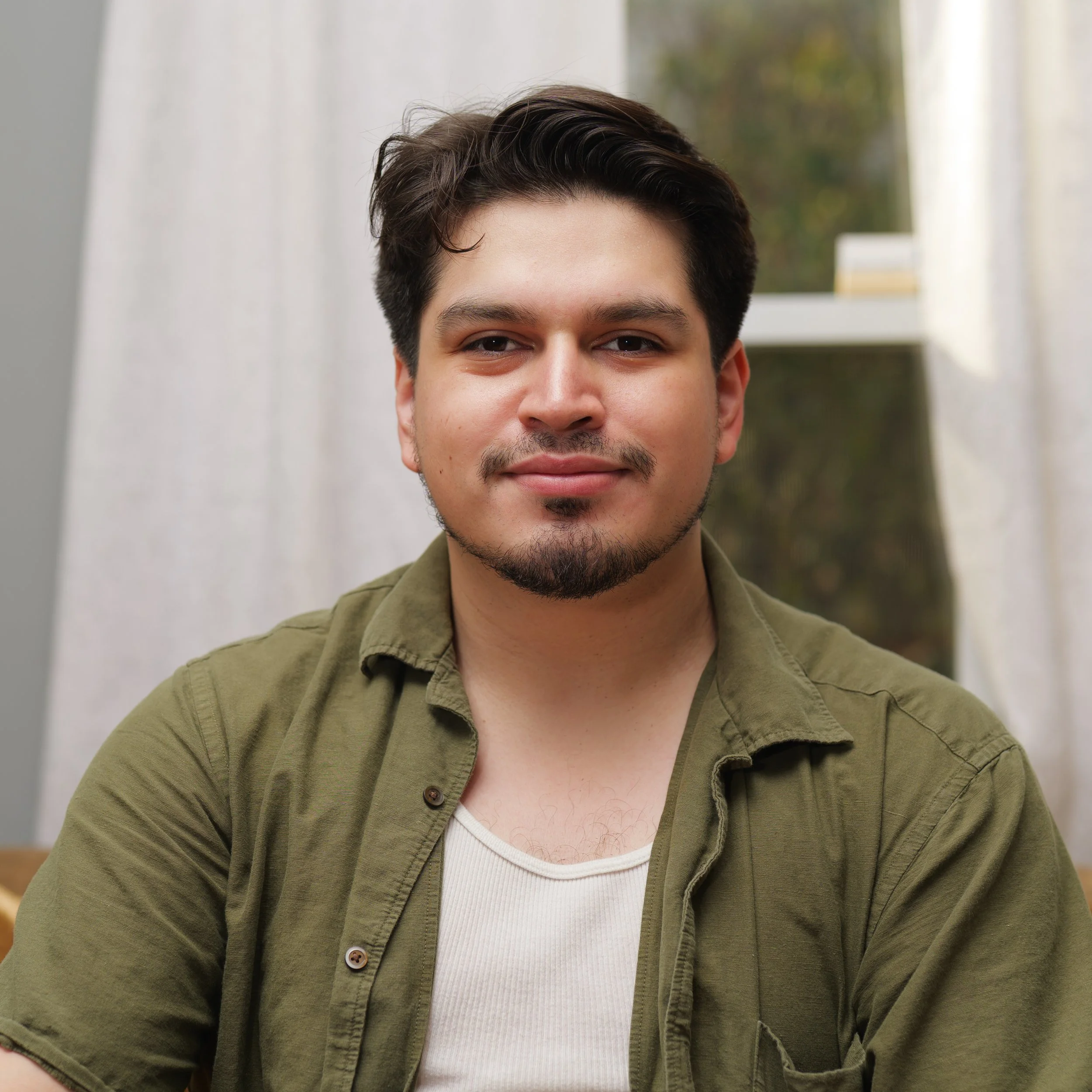 Close-up of a young man's face with dark hair, wearing a white shirt and an olive green jacket, smiling slightly inside a room with white curtains and a window showing trees outside.
