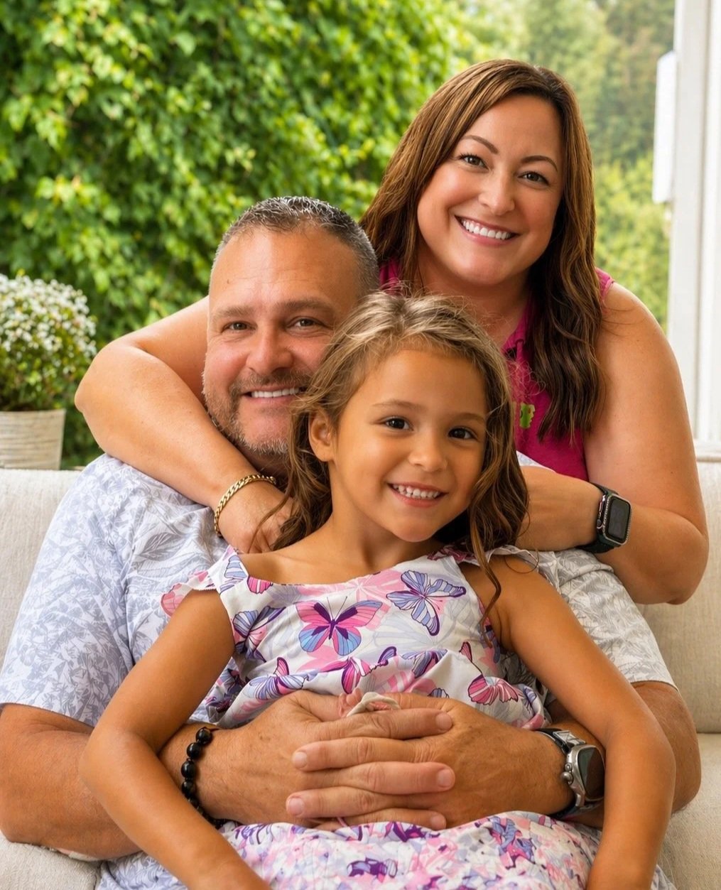 A smiling family of three sitting on a couch: a man with short hair and a beard, a woman with long brown hair, and a young girl with long curly hair. The woman is hugging the man from behind, and the girl is sitting in the man's lap. They are outside with greenery in the background.