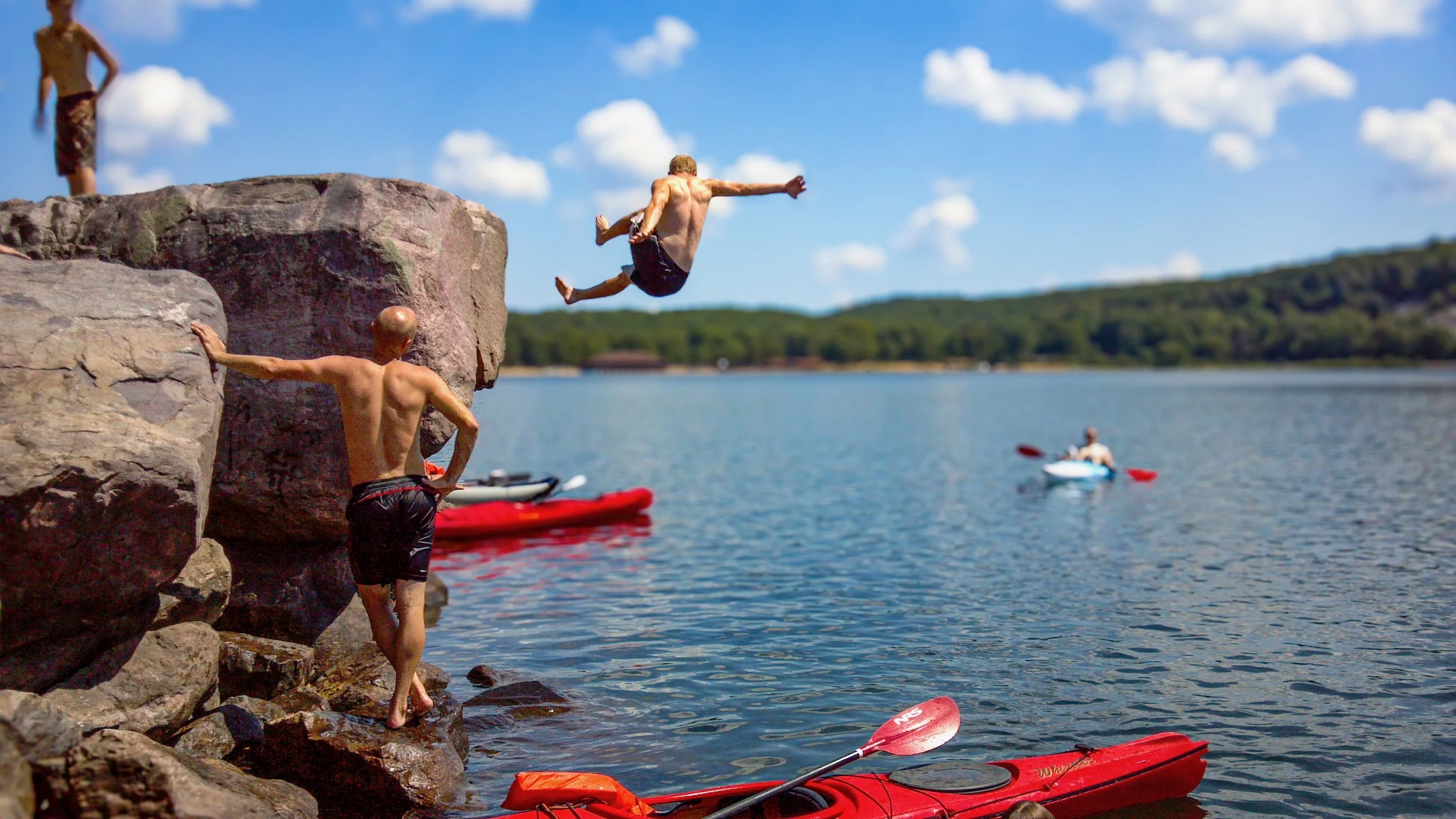 People jumping off rocks into a lake with kayaks and a person kayaking in the background, under a blue sky with clouds.