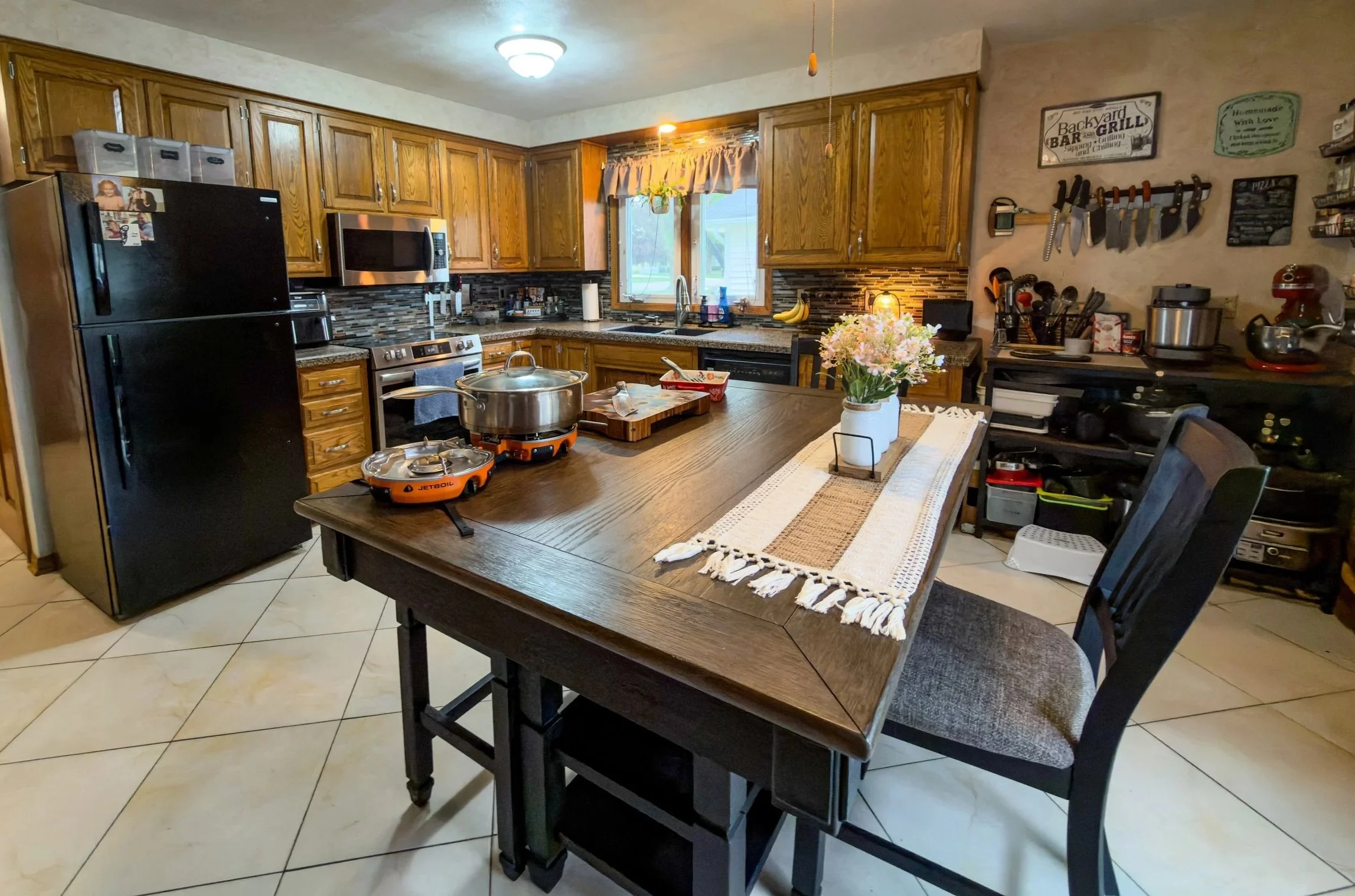 A kitchen with wooden cabinets, a black refrigerator, a microwave, and a stove. There is a wooden table with a flower vase, cooking utensils, and a portable stove on it. The walls are decorated with signs and hooks holding knives and kitchen tools. A window above the sink allows natural light into the room.