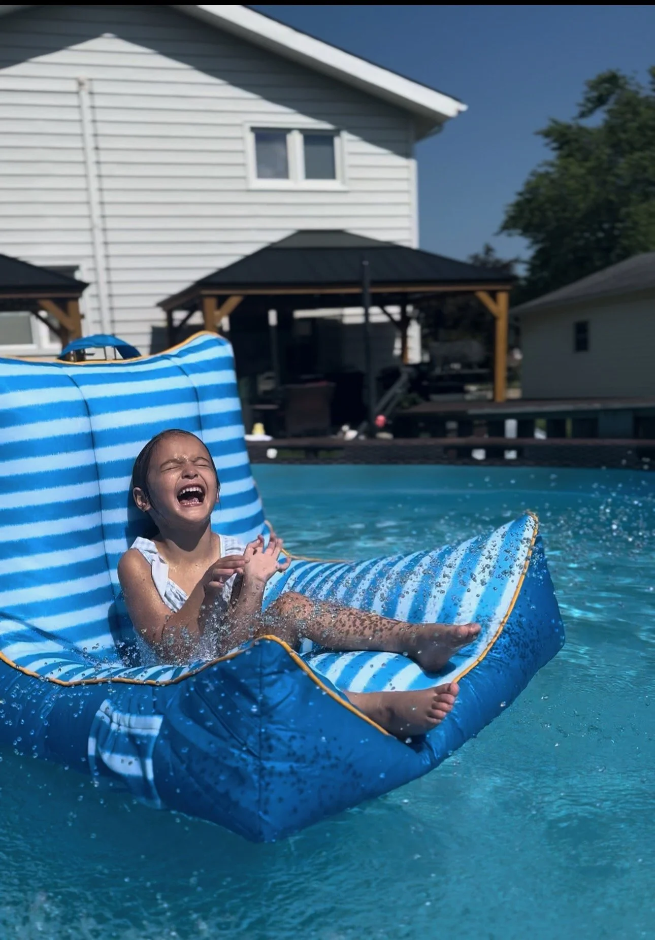 A young girl laughing and splashing water while sitting on an inflatable blue and white striped float in a backyard pool.