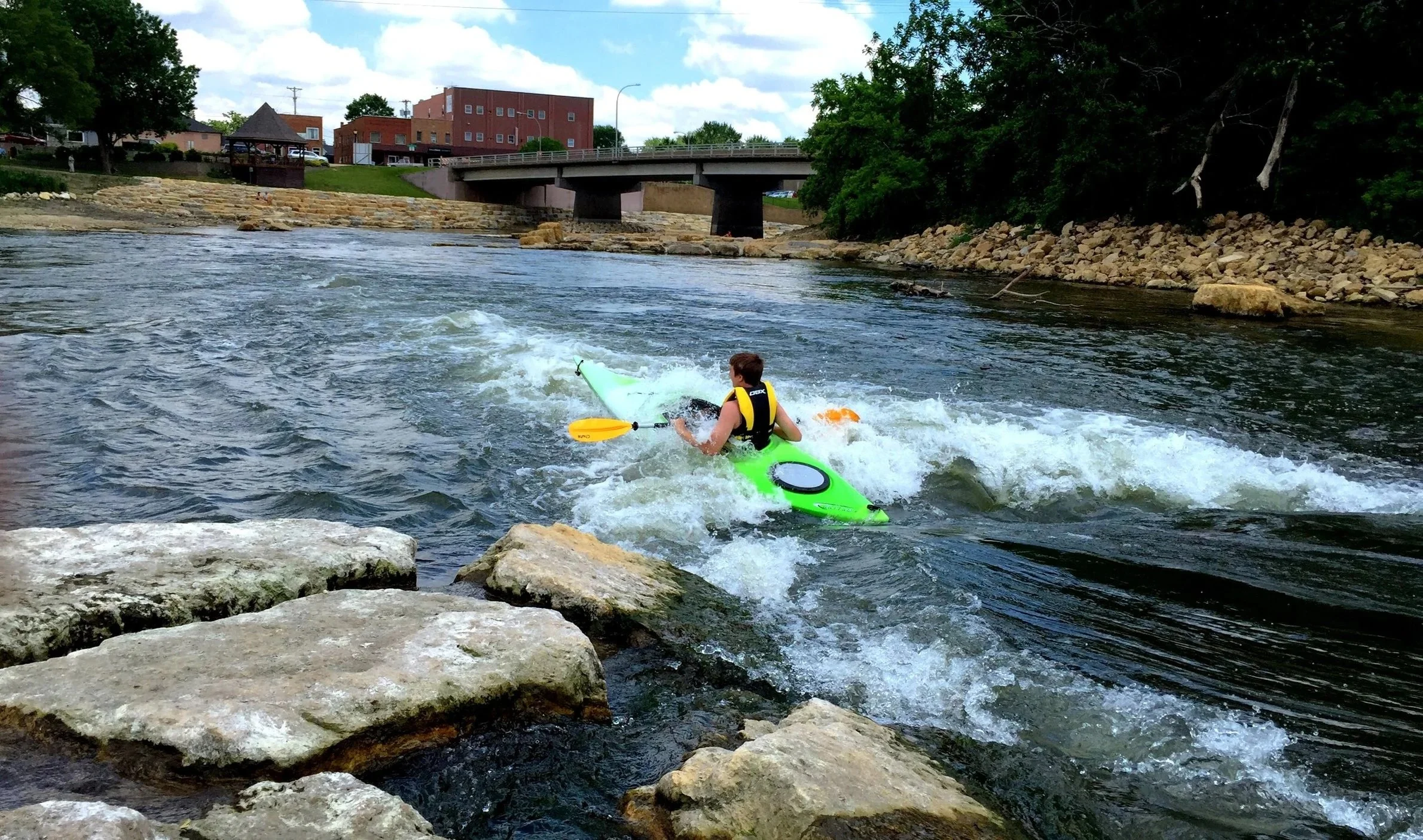 A person in a yellow life jacket kayaking on a river with rocks and trees on the sides, and a bridge and buildings in the background.