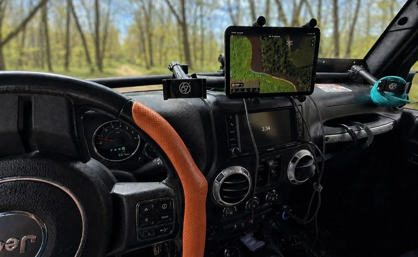 Interior of a vehicle dashboard with a Garmin GPS, a tablet mounted showing a map, and various controls, set against a background of trees visible through the windshield.