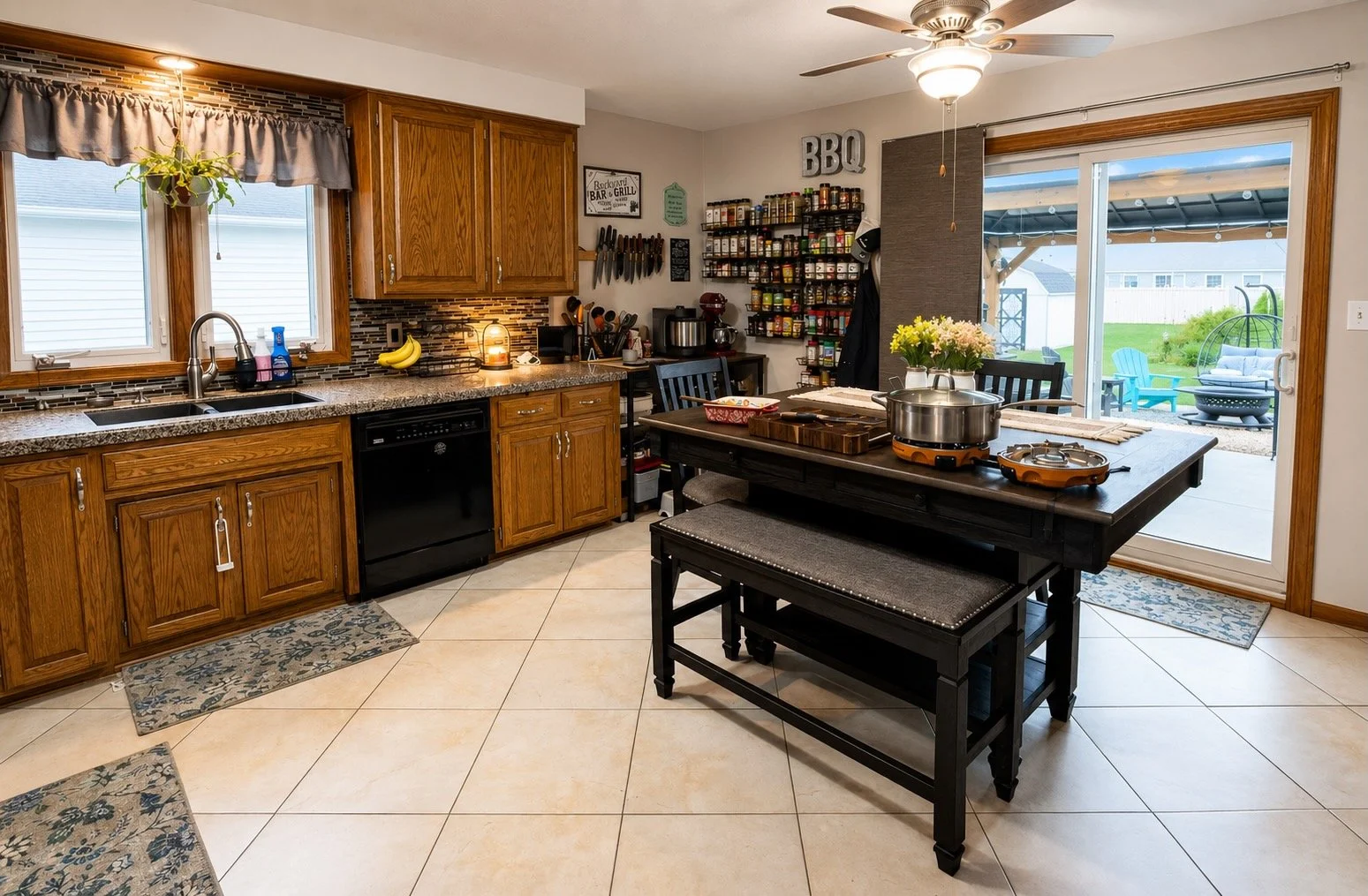 A kitchen with wooden cabinets, a tile backsplash, a window with a beige valance, a black dishwasher, and a countertop with bananas and soap. There is a dining table with a pot and flowers, and a sliding glass door leading outside to a patio with chairs and a fire pit.