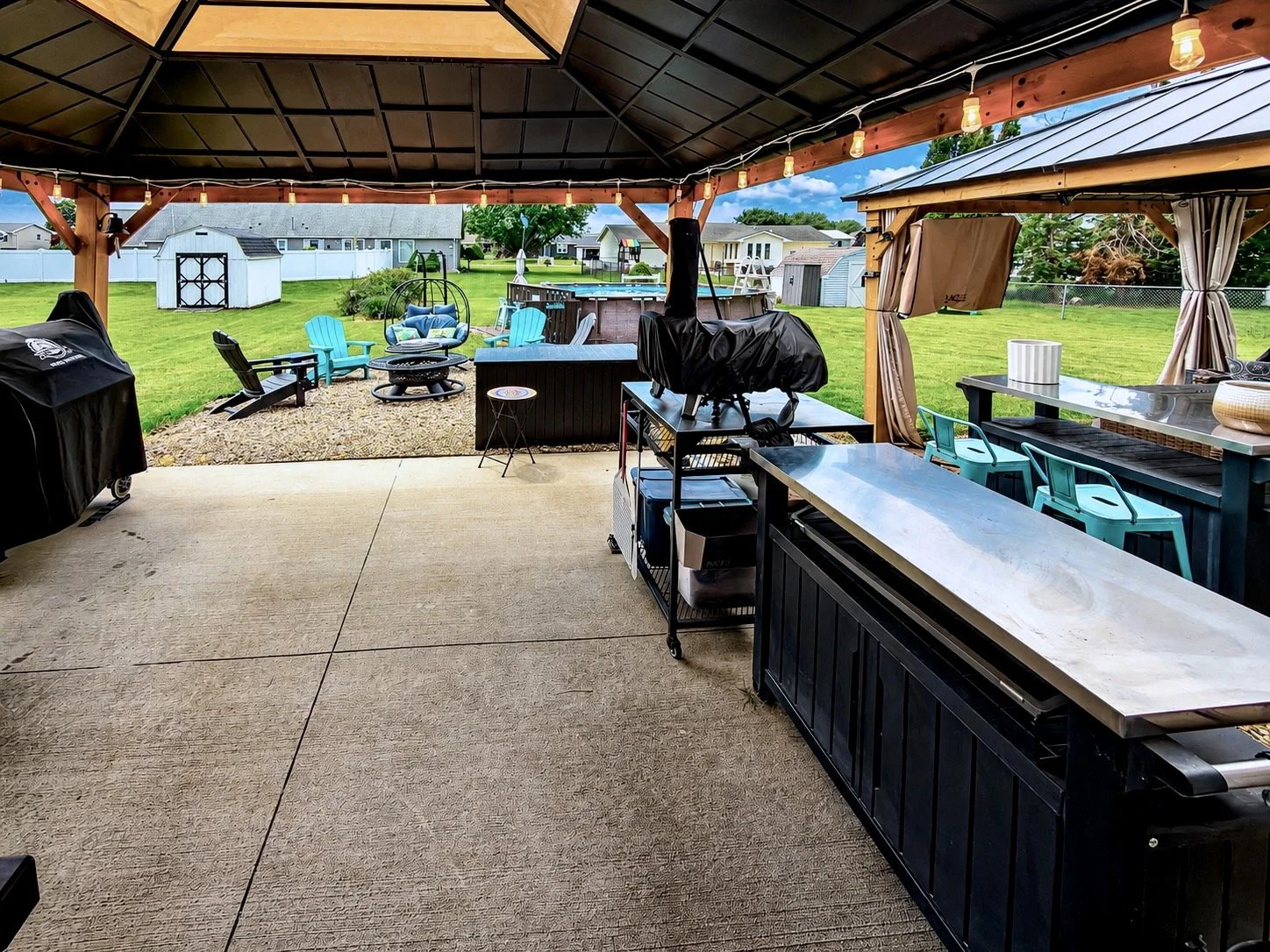 Backyard outdoor living space with covered patio, outdoor fireplace, lawn chairs, a shed, and a raised hot tub, with a view of open grassy yard and houses in the background.