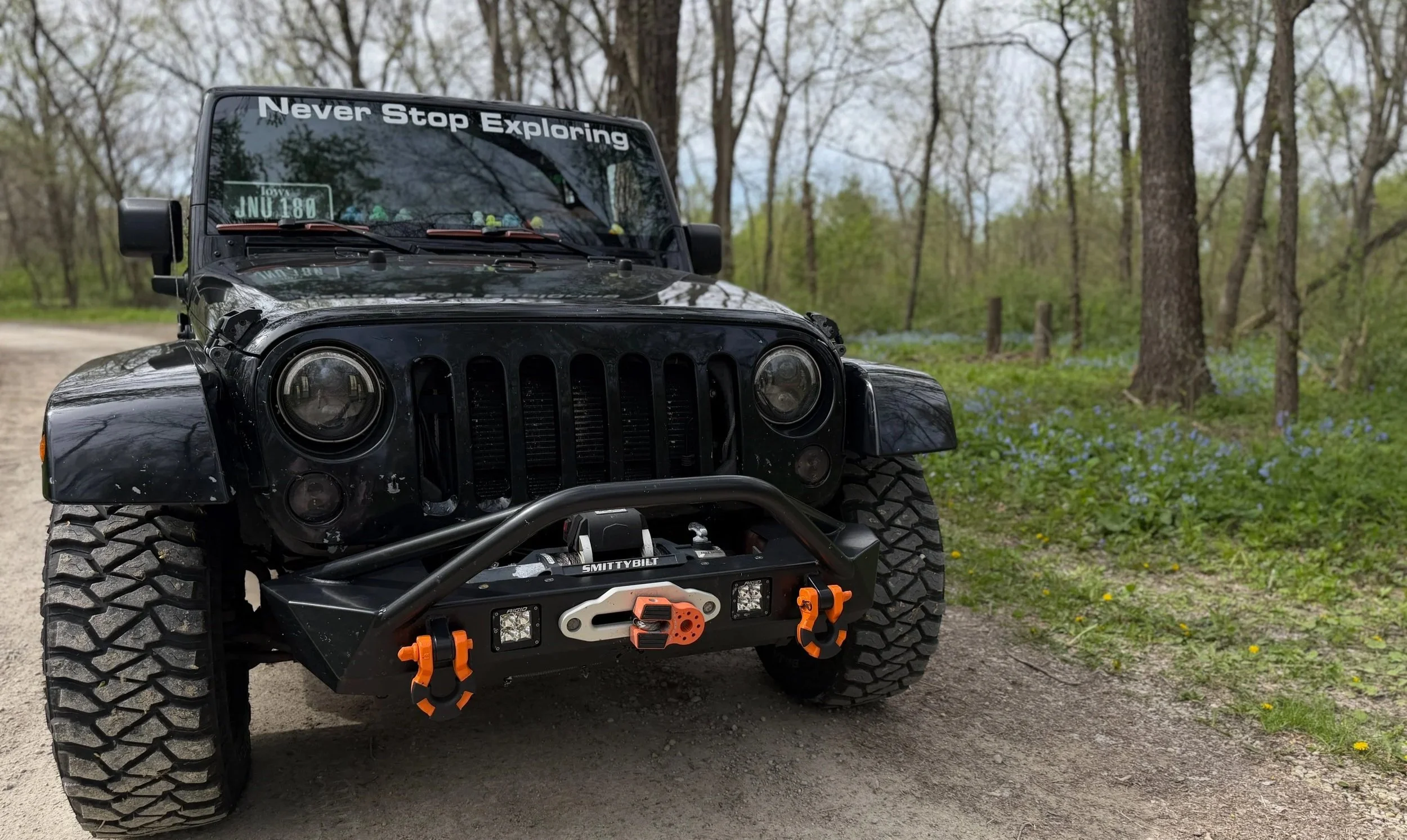 Close-up of black off-road vehicle with large tires, front bumper with orange hooks, and a windshield sign that says 'Never Stop Exploring', parked on a dirt path in a wooded area.