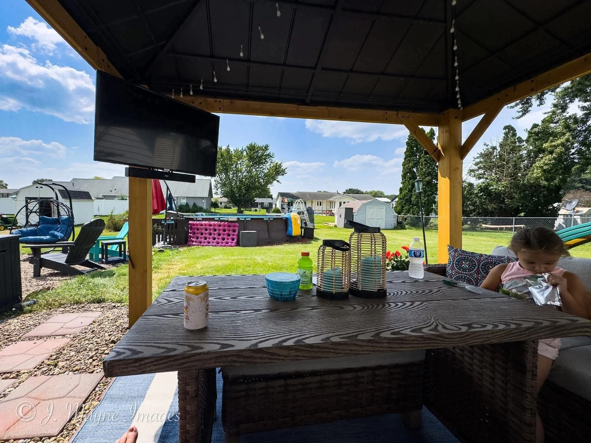 Backyard patio with a covered pergola, outdoor furniture, a TV, and children's play area under a partly cloudy sky.