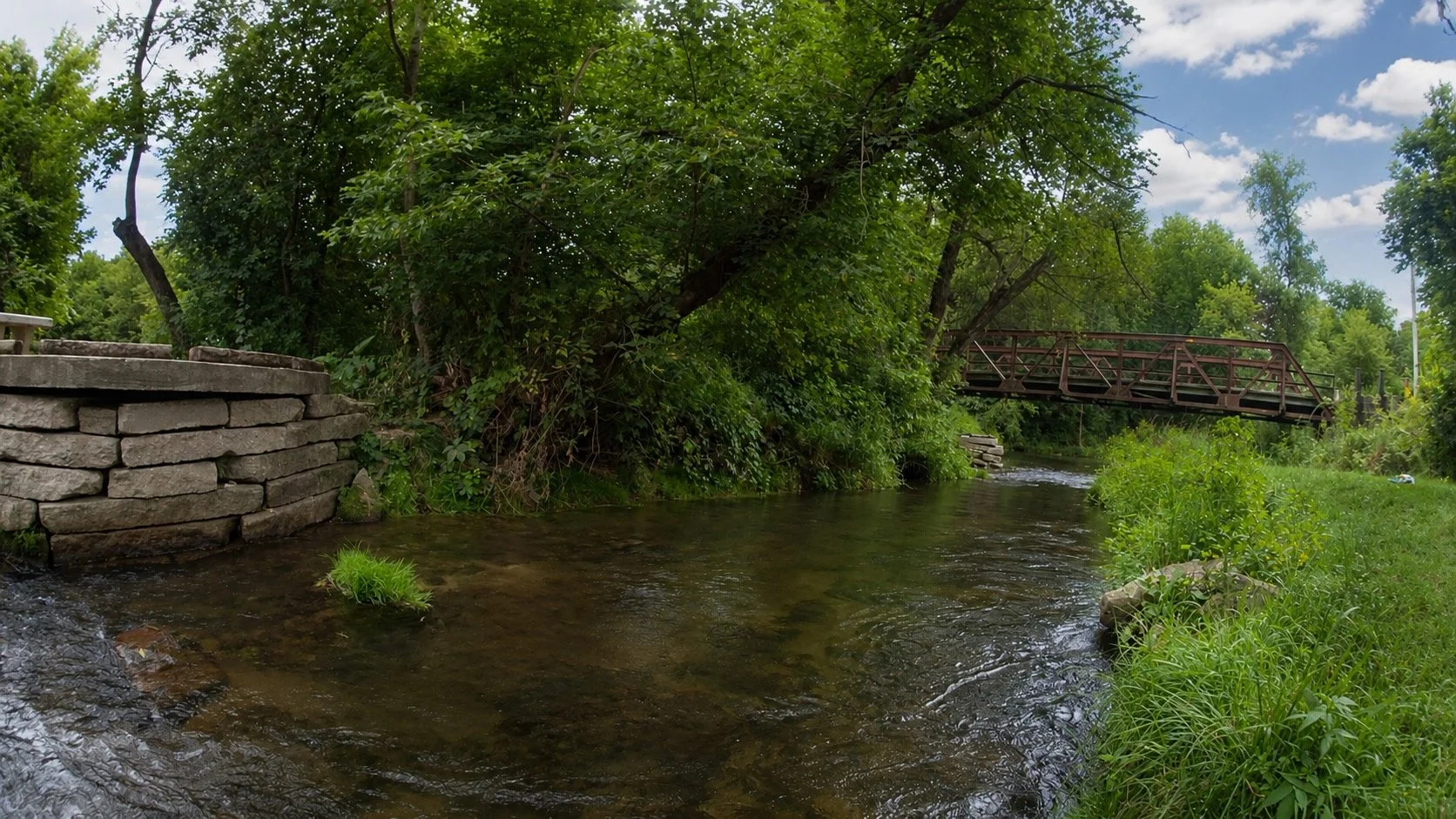A small stream with clear water flowing through a green, leafy area. There is a stone retaining wall on the left and a brown metal bridge in the background crossing over the stream. The sky is partly cloudy with blue patches visible.