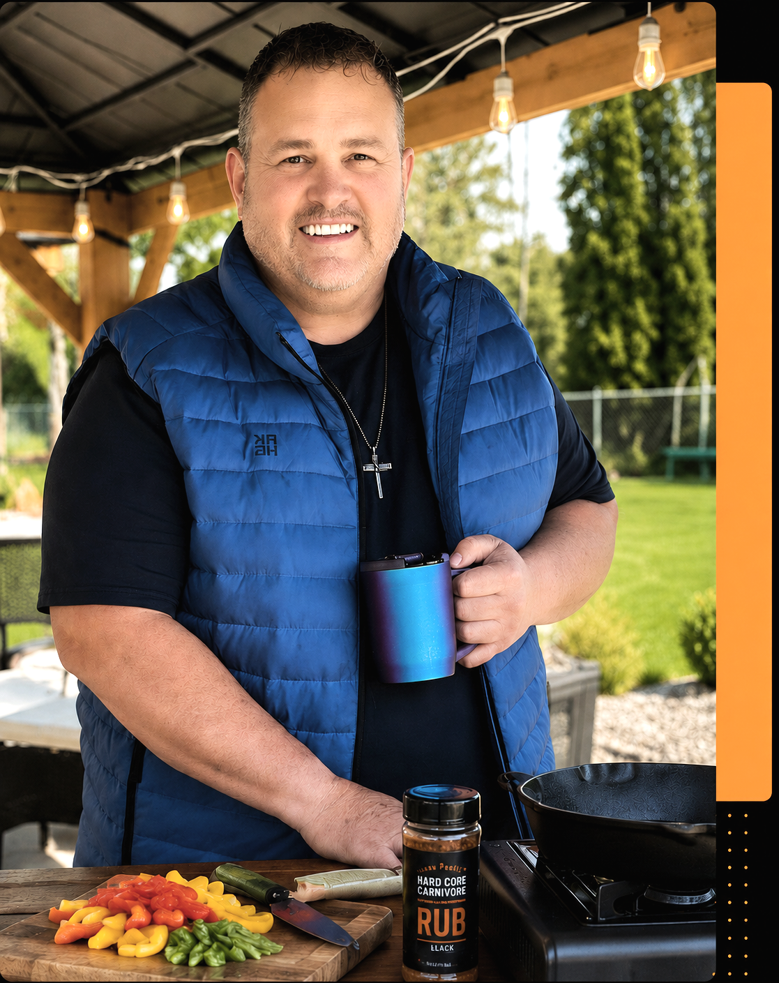 A smiling man in a blue vest and black shirt, holding a blue mug, standing at an outdoor cooking area with sliced vegetables, a seasoning jar, and a frying pan on a portable stove, with a green backyard in the background.