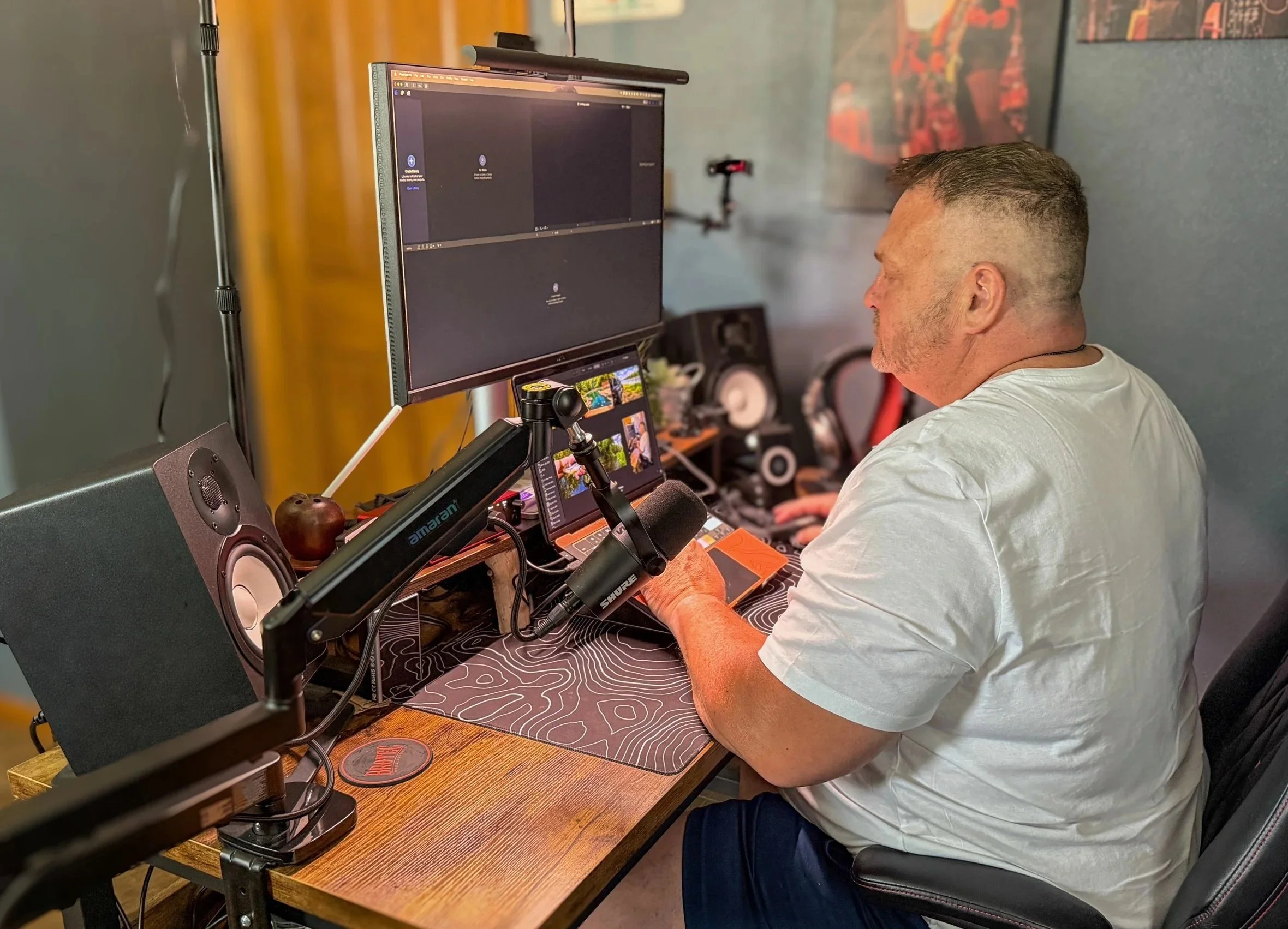 A man with short hair and a white t-shirt sitting at a desk in front of a large monitor. The desk has a computer mouse, mouse pad, and a phone. There are speakers, a camera, and other electronic equipment on the desk. The background has a wall with a colorful map or artwork.