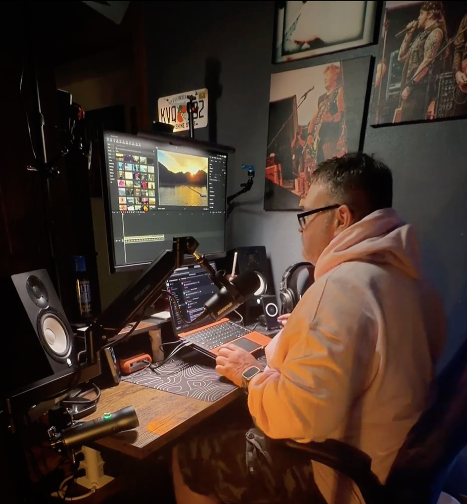 A person working on a computer in a dark room with music posters on the wall, a microphone attached to a desk arm, studio speakers, and a monitor showing a photo editing program.