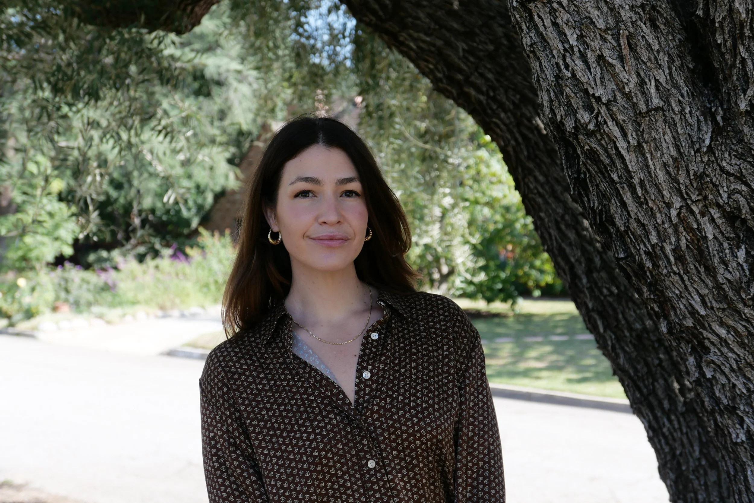 A woman standing outdoors beside a tree in a park, with greenery and flowers in the background.