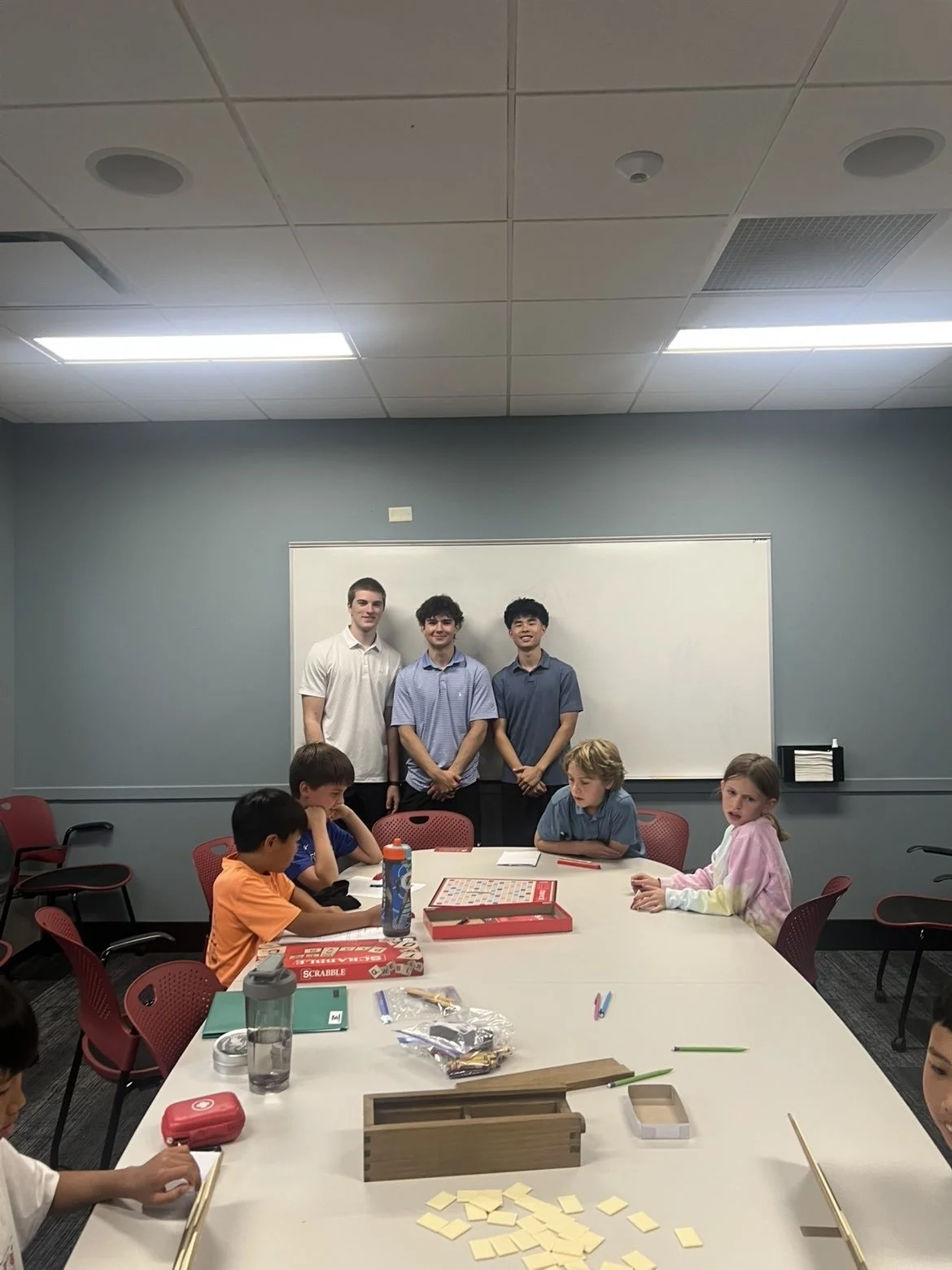 Group photo of six children, three standing in front of a whiteboard and three sitting at a table with game and school supplies in a classroom.