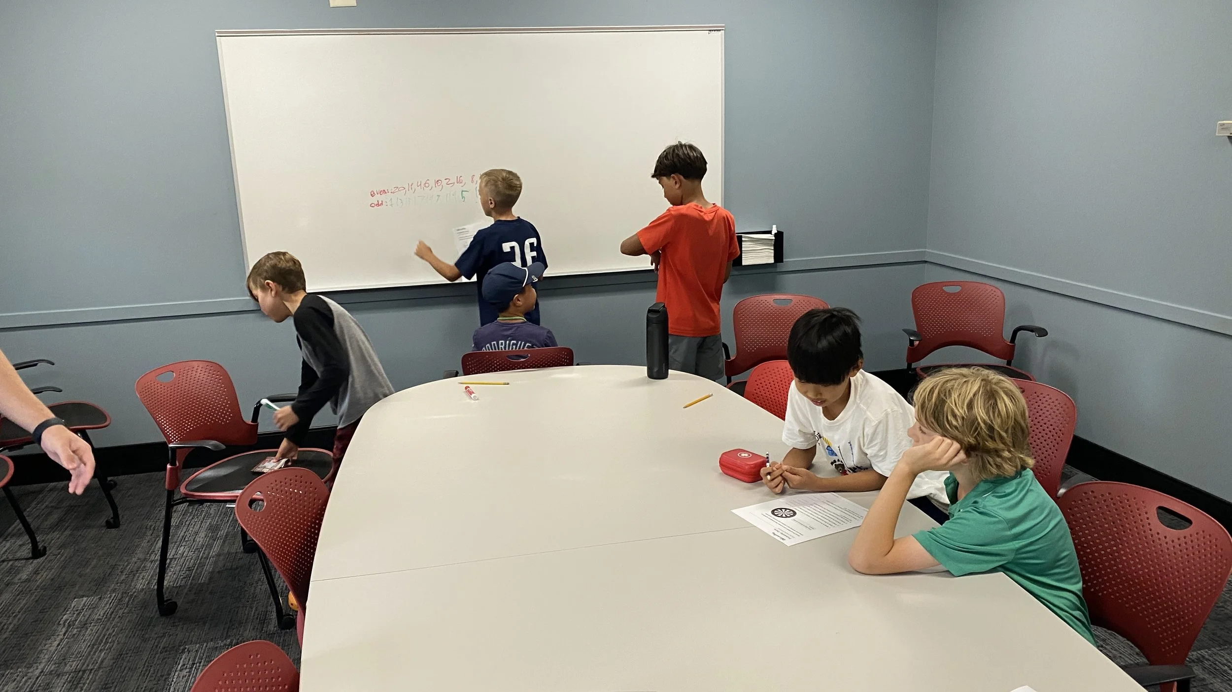 A classroom with children gathering around a whiteboard, some writing on the board, others sitting at a table with papers and supplies, in a room with gray walls and red chairs.