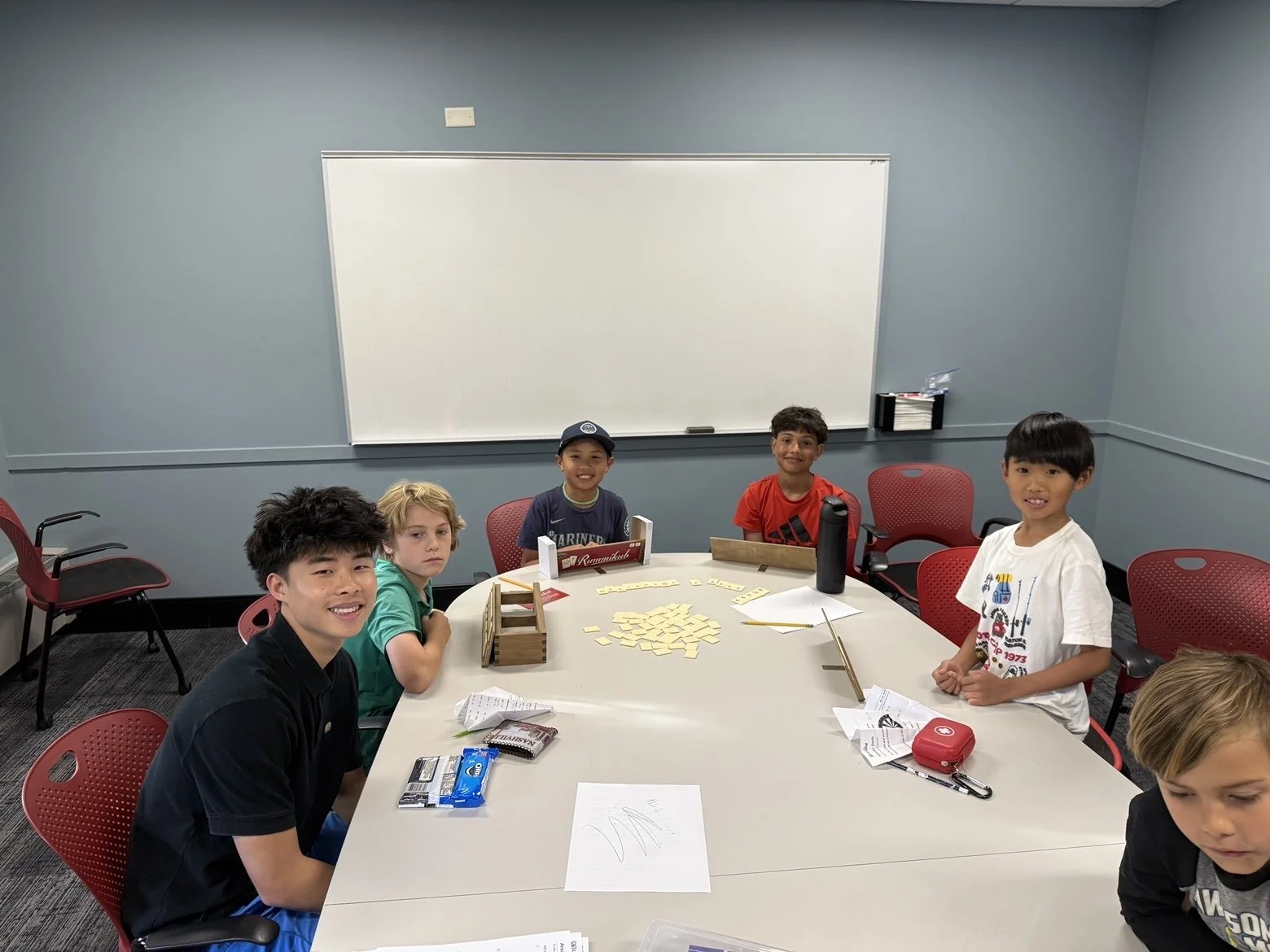 Six children sitting around a conference table in a classroom, smiling at the camera. The table has game pieces, papers, and snacks on it; a whiteboard is in the background.