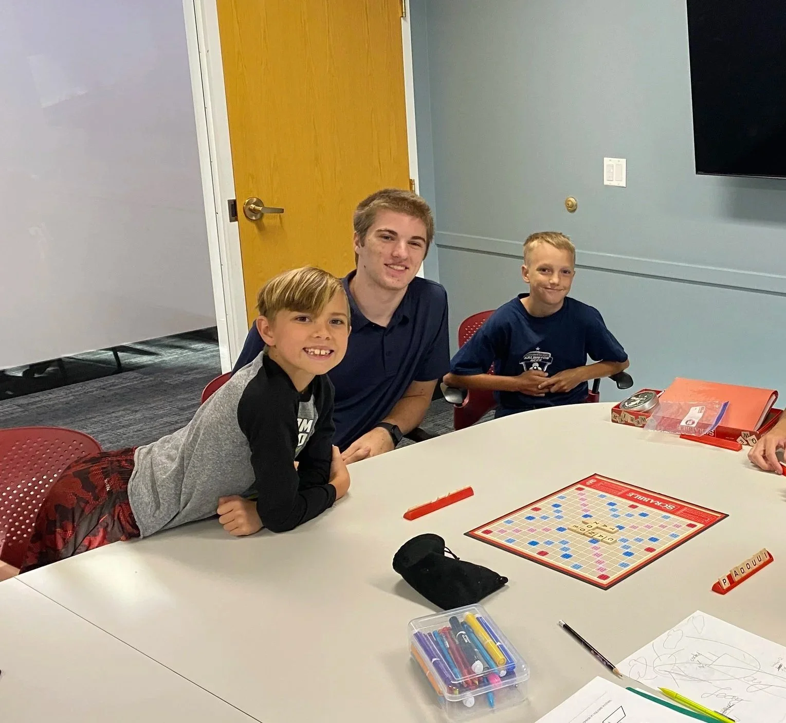 Three children sitting at a table playing Scrabble, with game pieces and markers on the table, in a room with a yellow door and a gray wall.