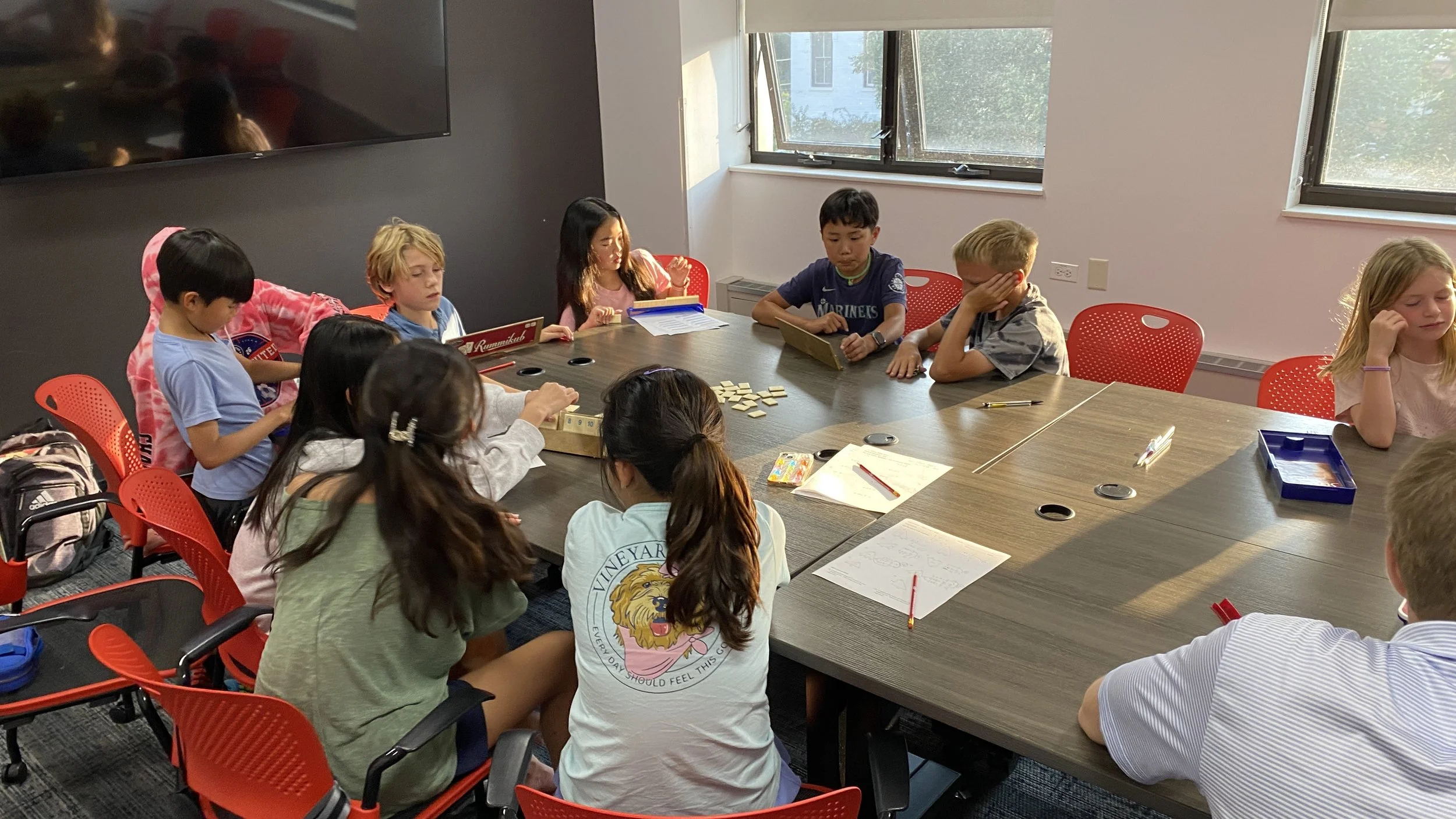 Children sitting around a conference table engaged in an activity, with some playing a game and others writing or reading, in a brightly lit room with windows.