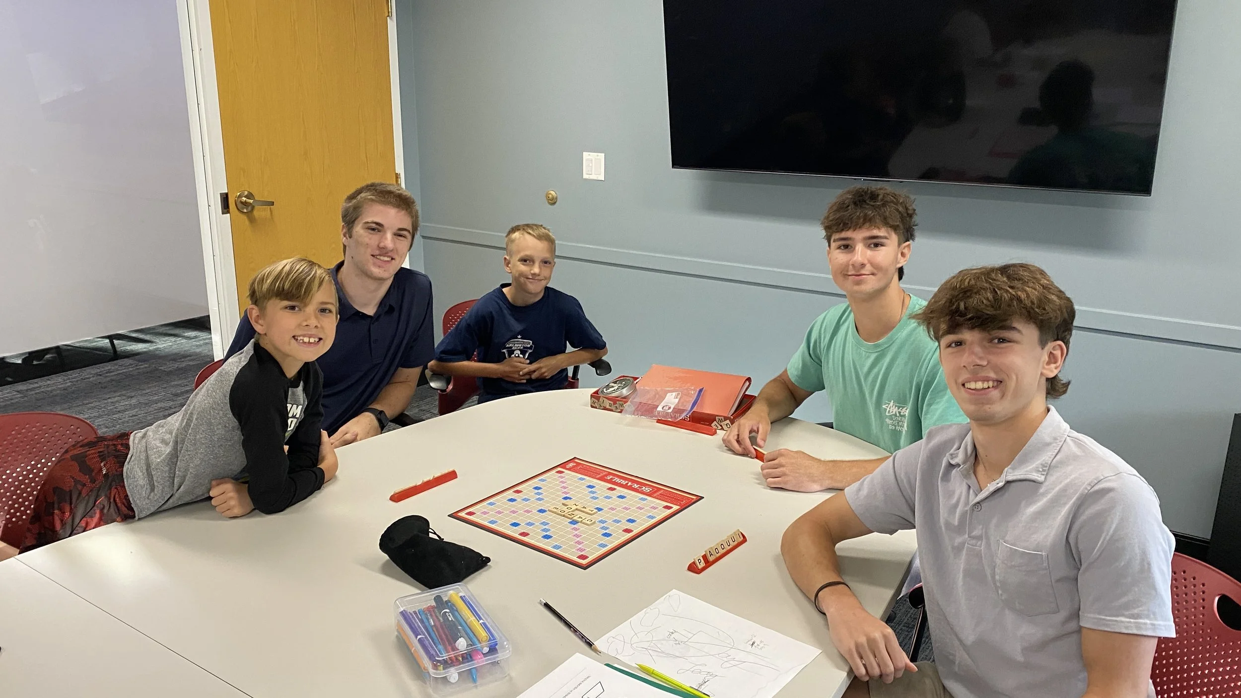 A group of five boys sitting around a white table playing Scrabble in a room with light blue walls and a large black television mounted on the wall. The boys are smiling and have various items on the table, including game pieces, markers, and papers.