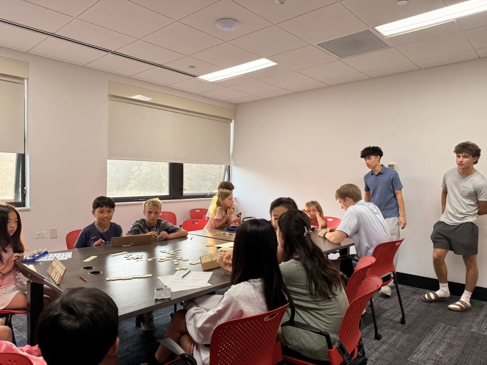 A group of children and teenagers gathered around a large conference table in a classroom or meeting room, some sitting and some standing, with various games or activities spread across the table. The room has white walls, large windows with blinds, 