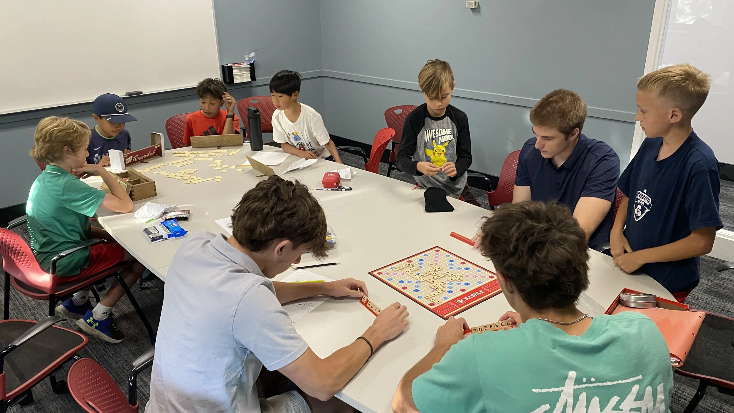 Group of boys and young men playing Scrabble and other board games around a large conference table in a classroom or game room.
