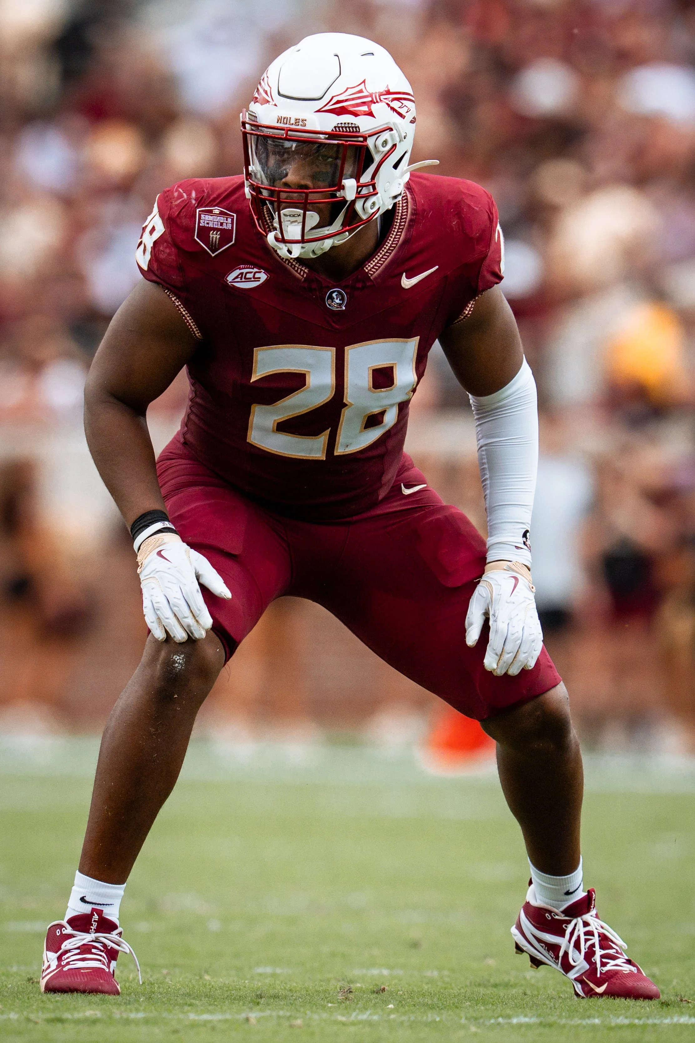 An American football player in red uniform with the number 28, crouched in a ready stance on the field, wearing a white helmet with red graphics, white gloves, and red and white shoes.