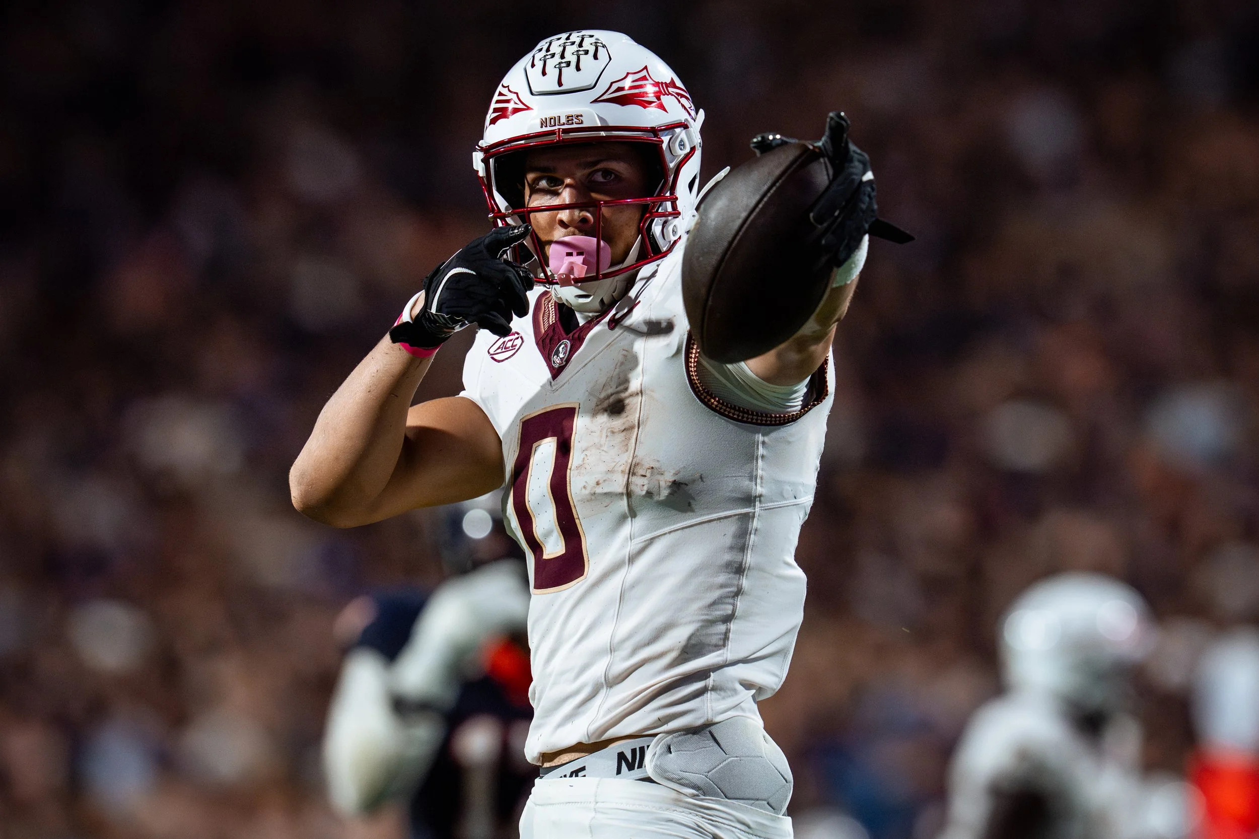 A football player wearing a white uniform with maroon and gold accents, a helmet with a maroon and white design, and a pink mouthguard, is pointing towards the camera on the field.
