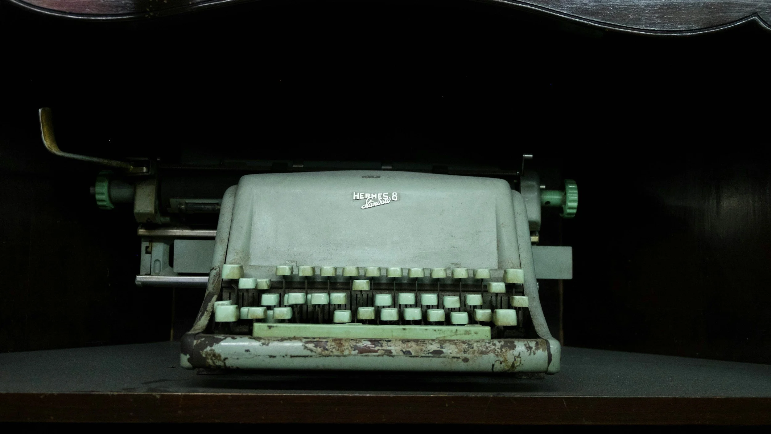 An old, vintage Hermes 8 typewriter with faded green keys, placed on a dark surface in a dimly lit environment.