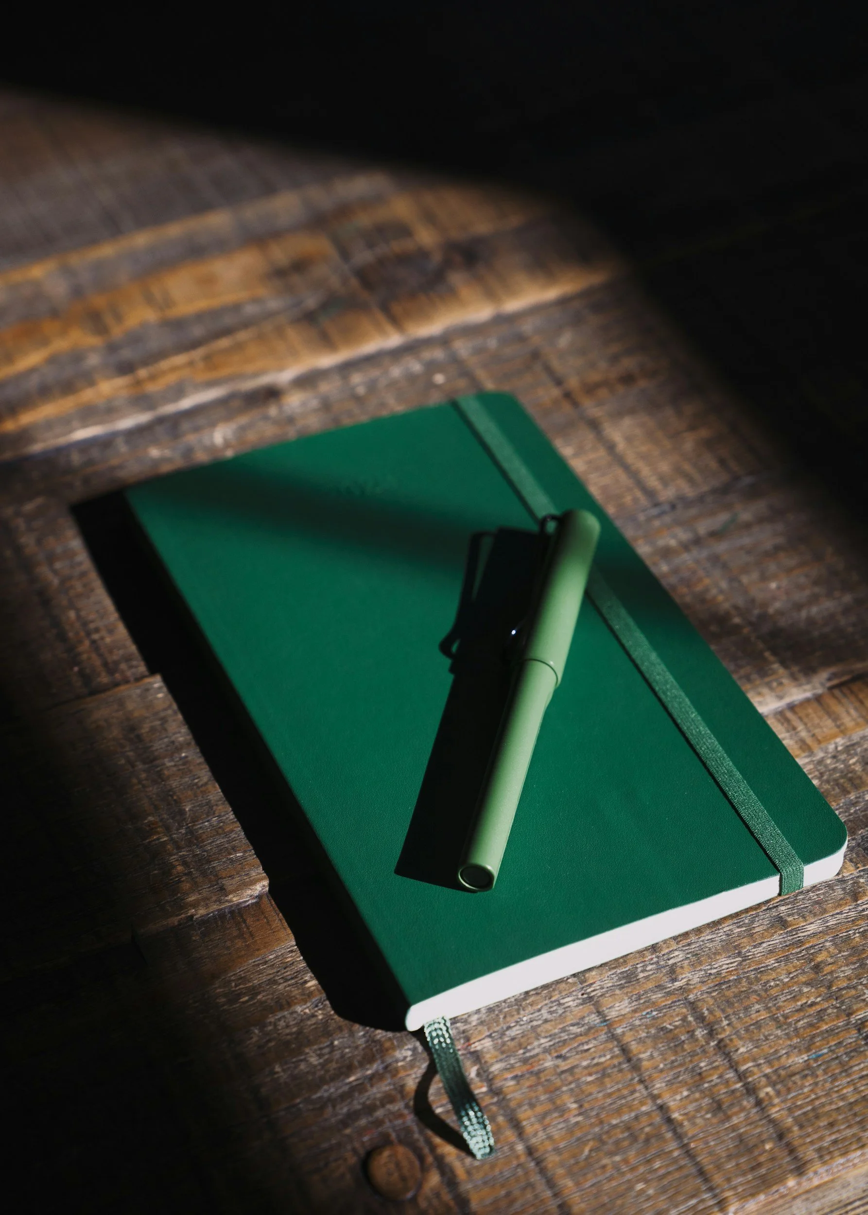 A green hardcover notebook with a pen placed on it, lying on a wooden surface.