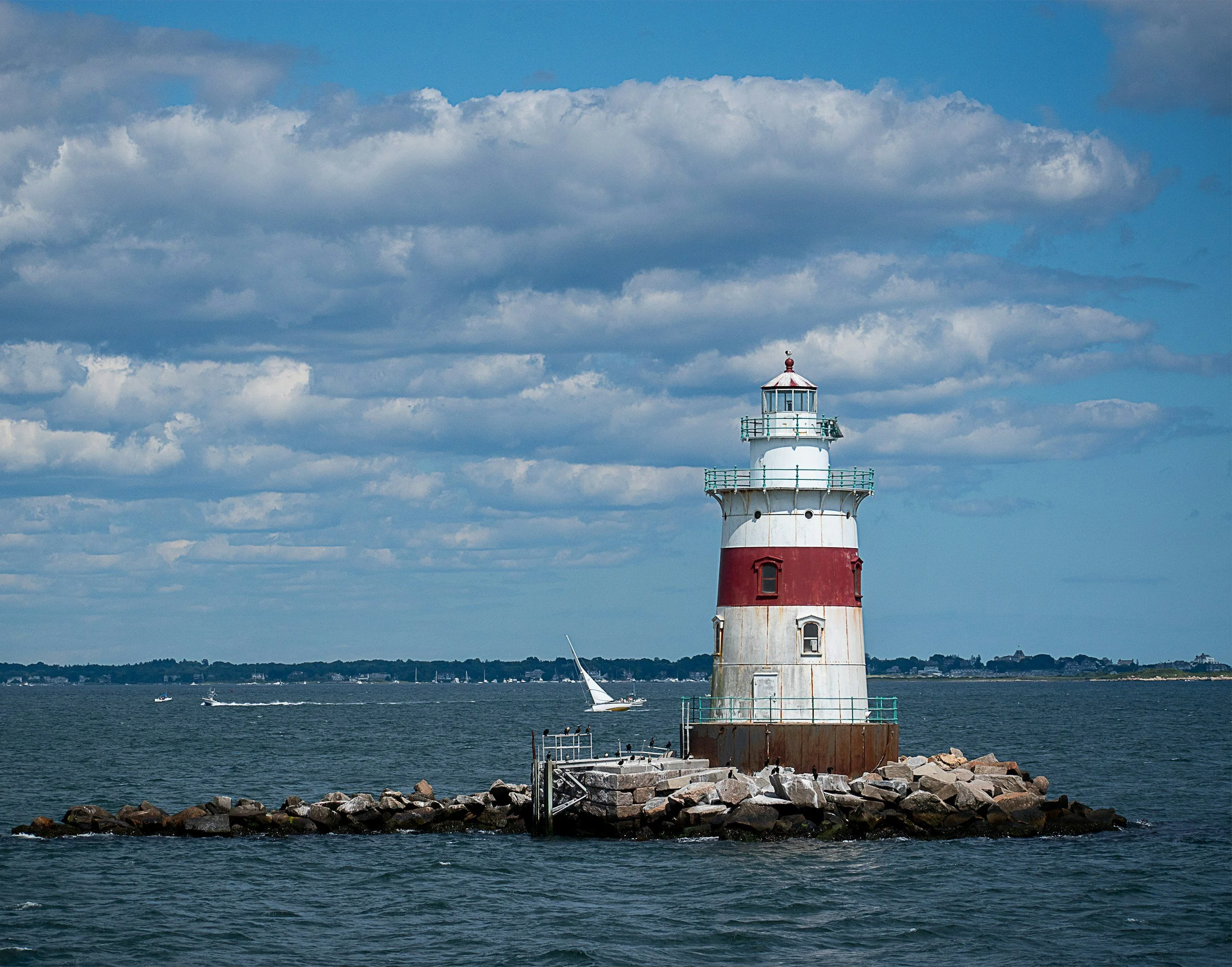A lighthouse standing on rocky shoreline with sailboats in the water, cloudy skies in the background.