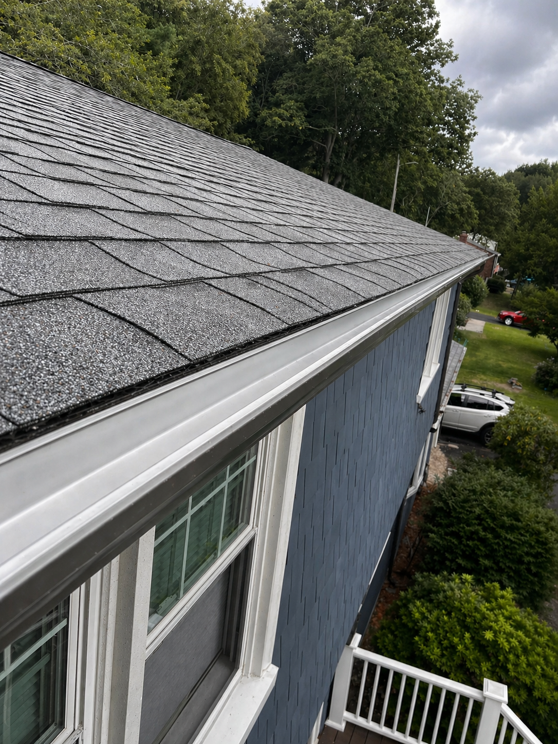 Wet gray asphalt shingle roof on blue house with white window frames and surrounded by trees and a green lawn.