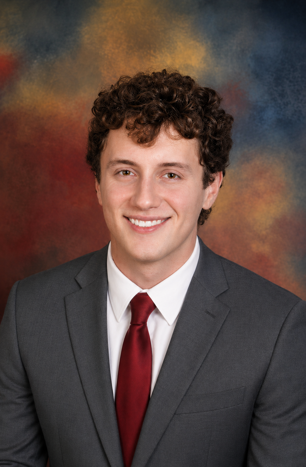 A young man with curly brown hair, wearing a dark gray suit, white shirt, and red tie, smiling in front of a colorful, abstract background.