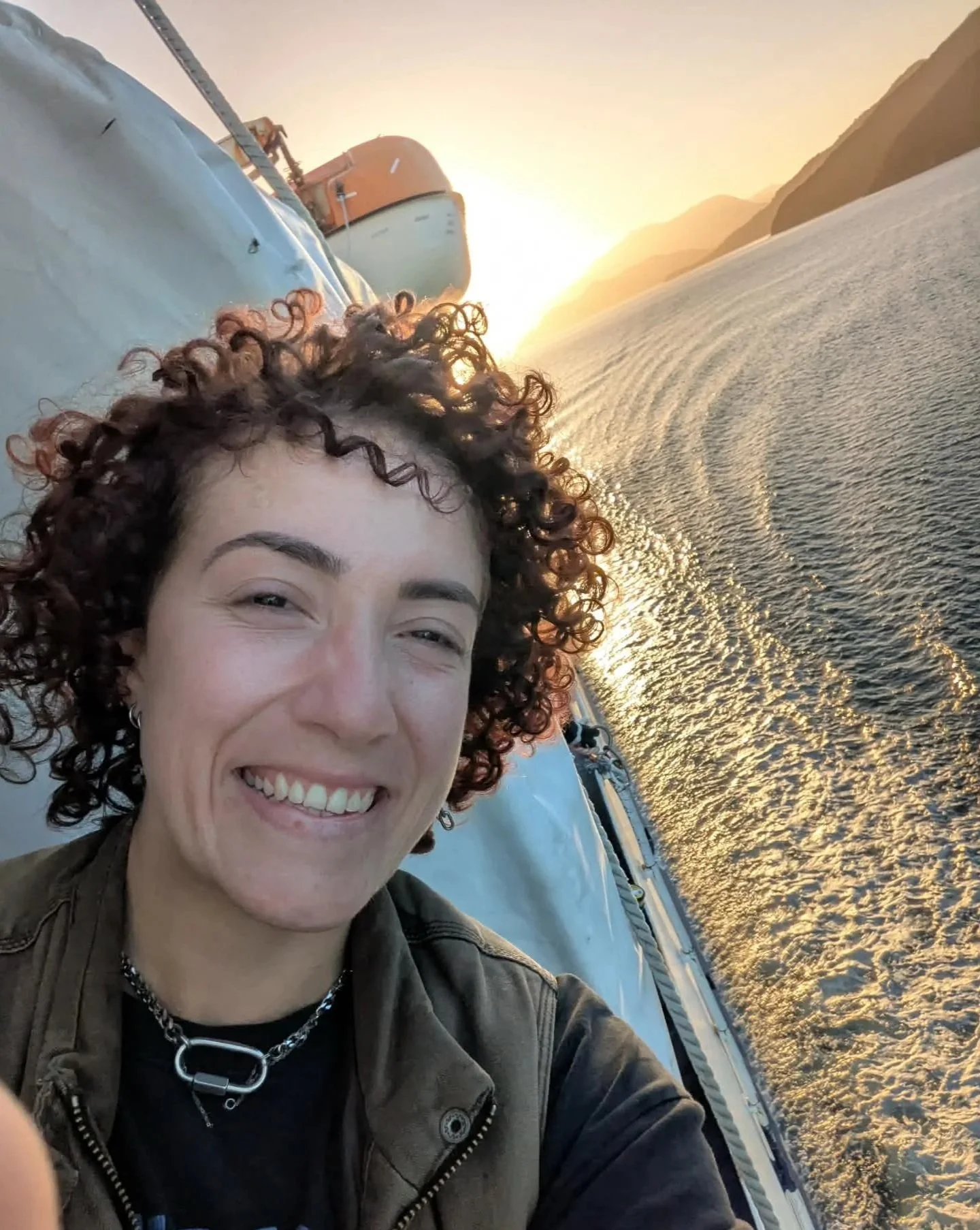 A woman with curly brown hair smiling on a boat at sunset with mountains in the background.