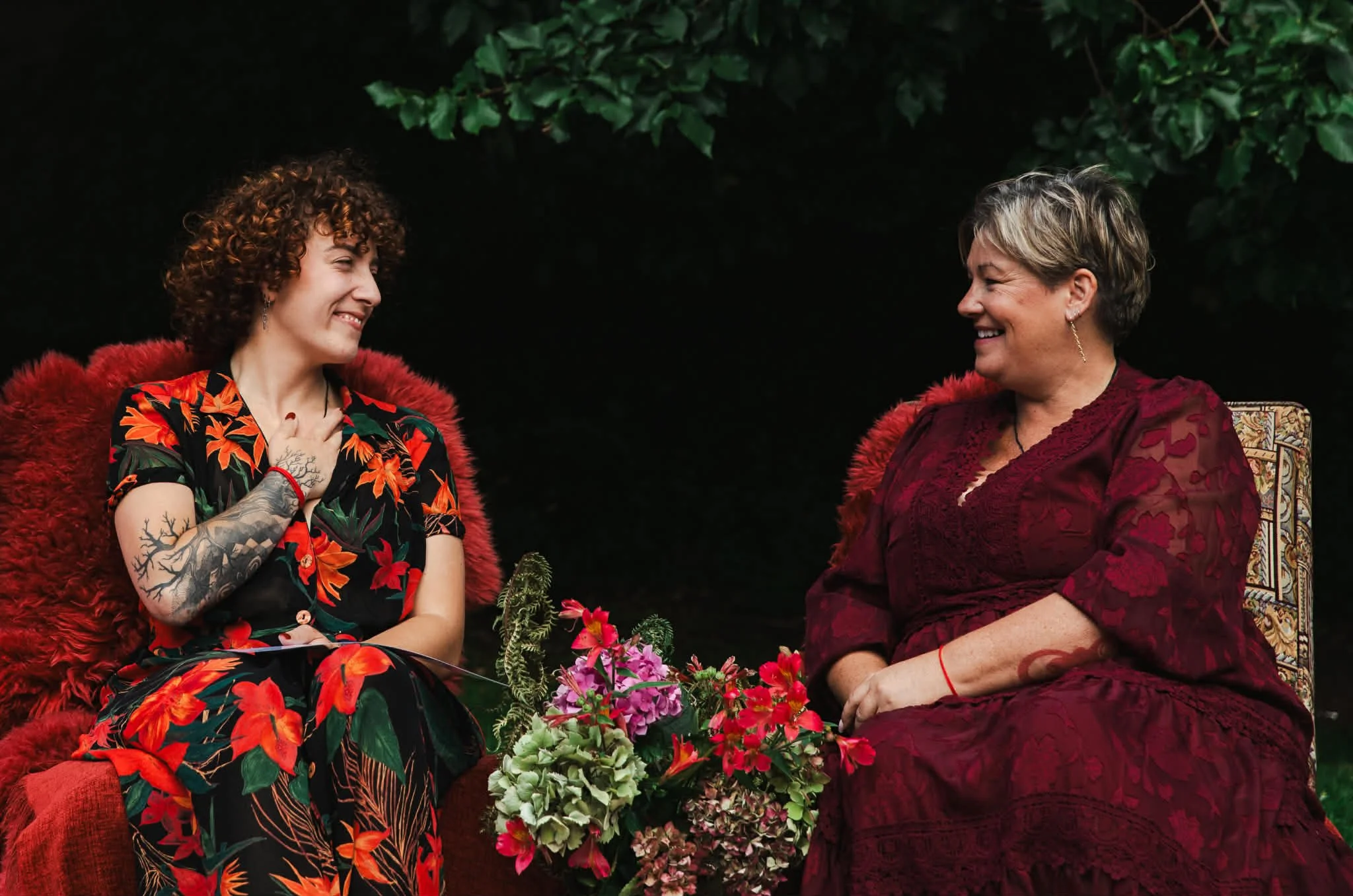 Two women sitting outdoors, smiling and engaging in conversation, surrounded by green foliage and flowers, with red and floral-patterned clothing.